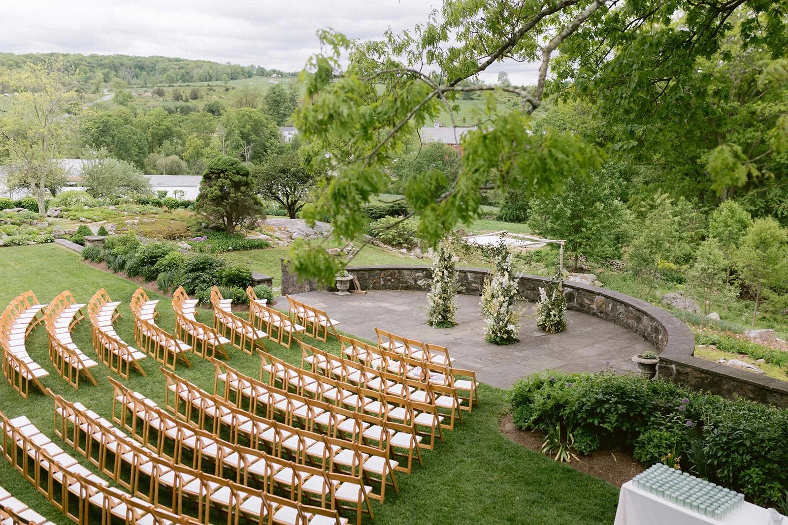Outdoor wedding ceremony setup with rows of wooden chairs on grass, white cushions, an altar with floral arrangements, surrounded by greenery and trees, hill in the background.