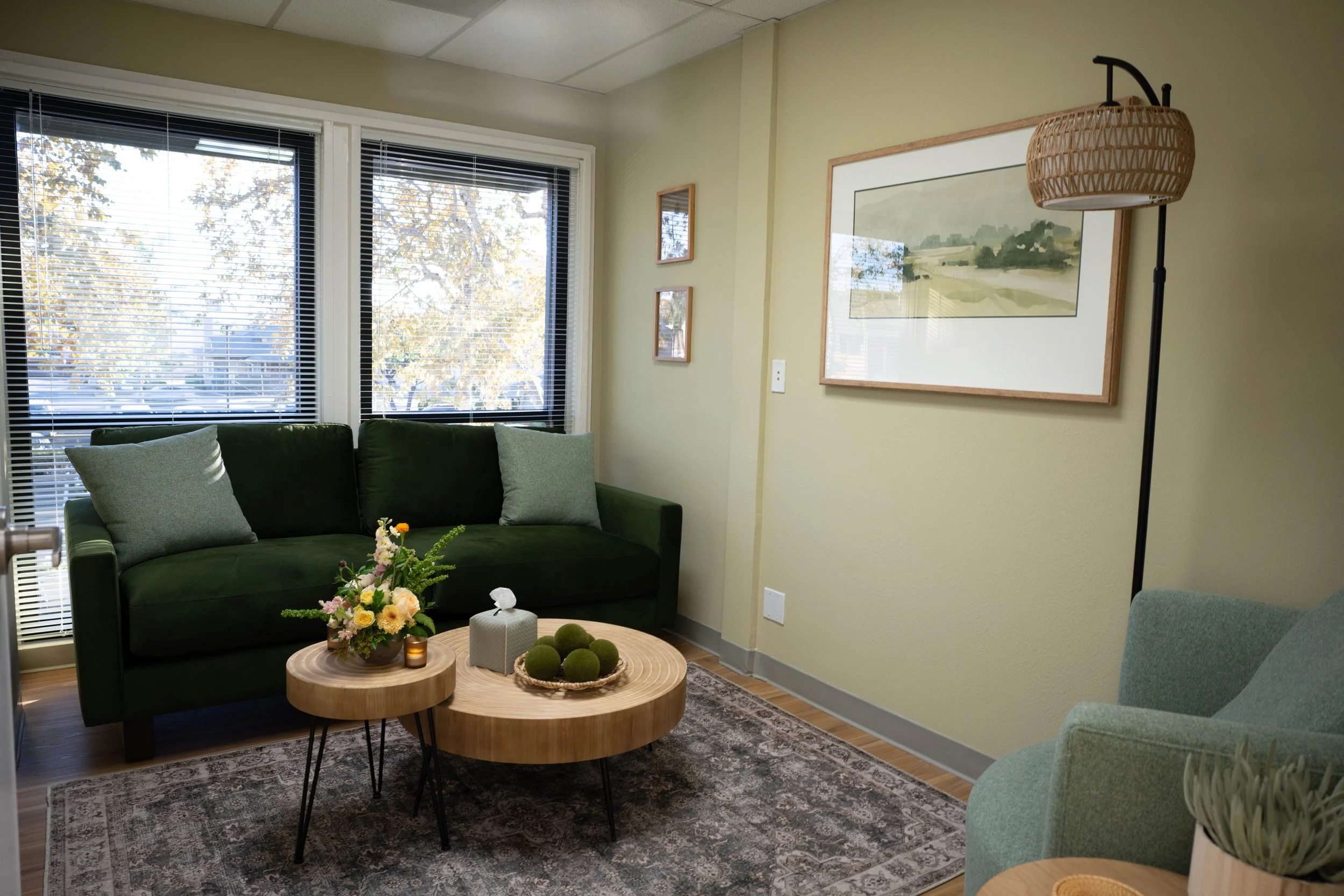 Cozy living room with a green sofa, light green armchair, wooden coffee tables, a floral arrangement, and wall art, illuminated by natural light through large windows with blinds.