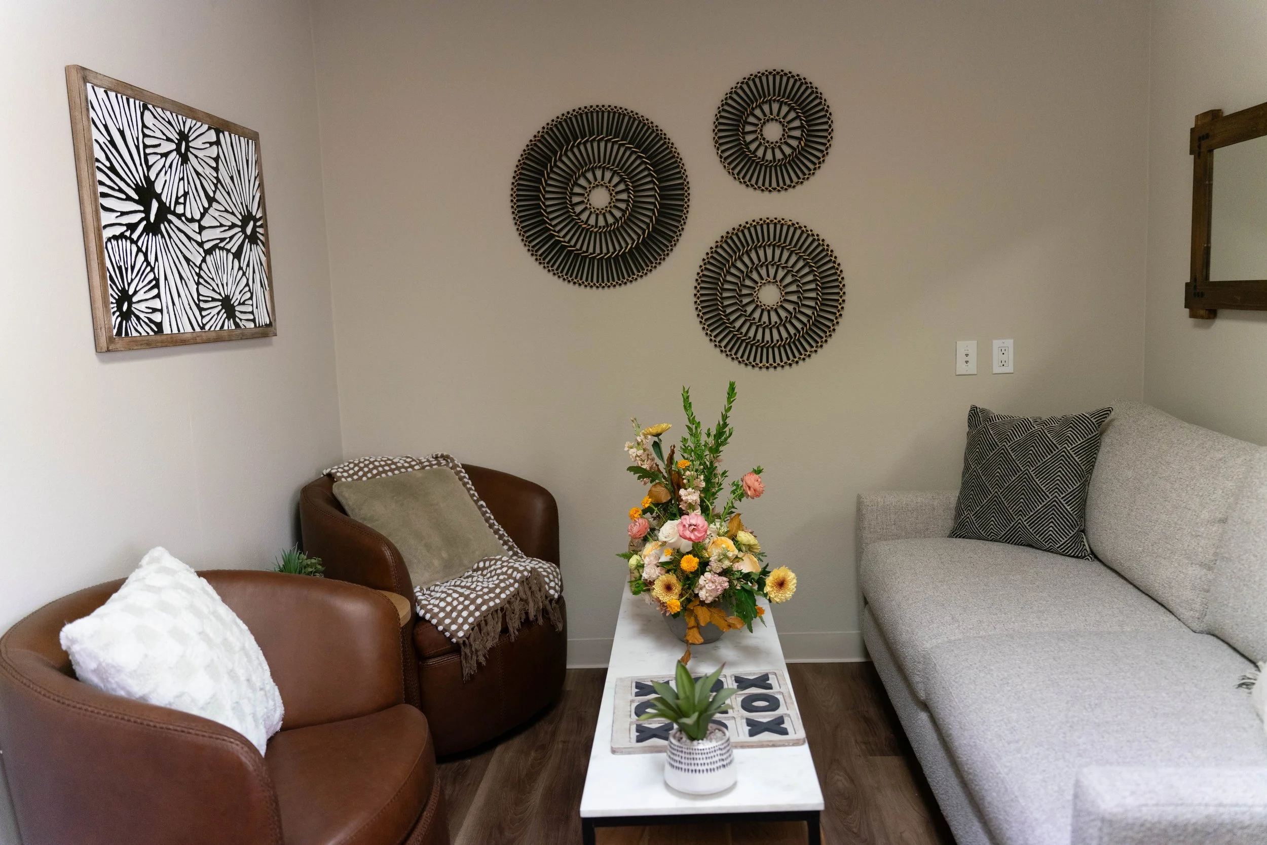 Living room with a gray couch, a brown leather chair, and a small armchair with a pillow. A white table with a floral arrangement and a small potted plant is in the center. Decorative wall art includes three circular woven pieces and a framed black and white floral print. There is a mirror on the wall.