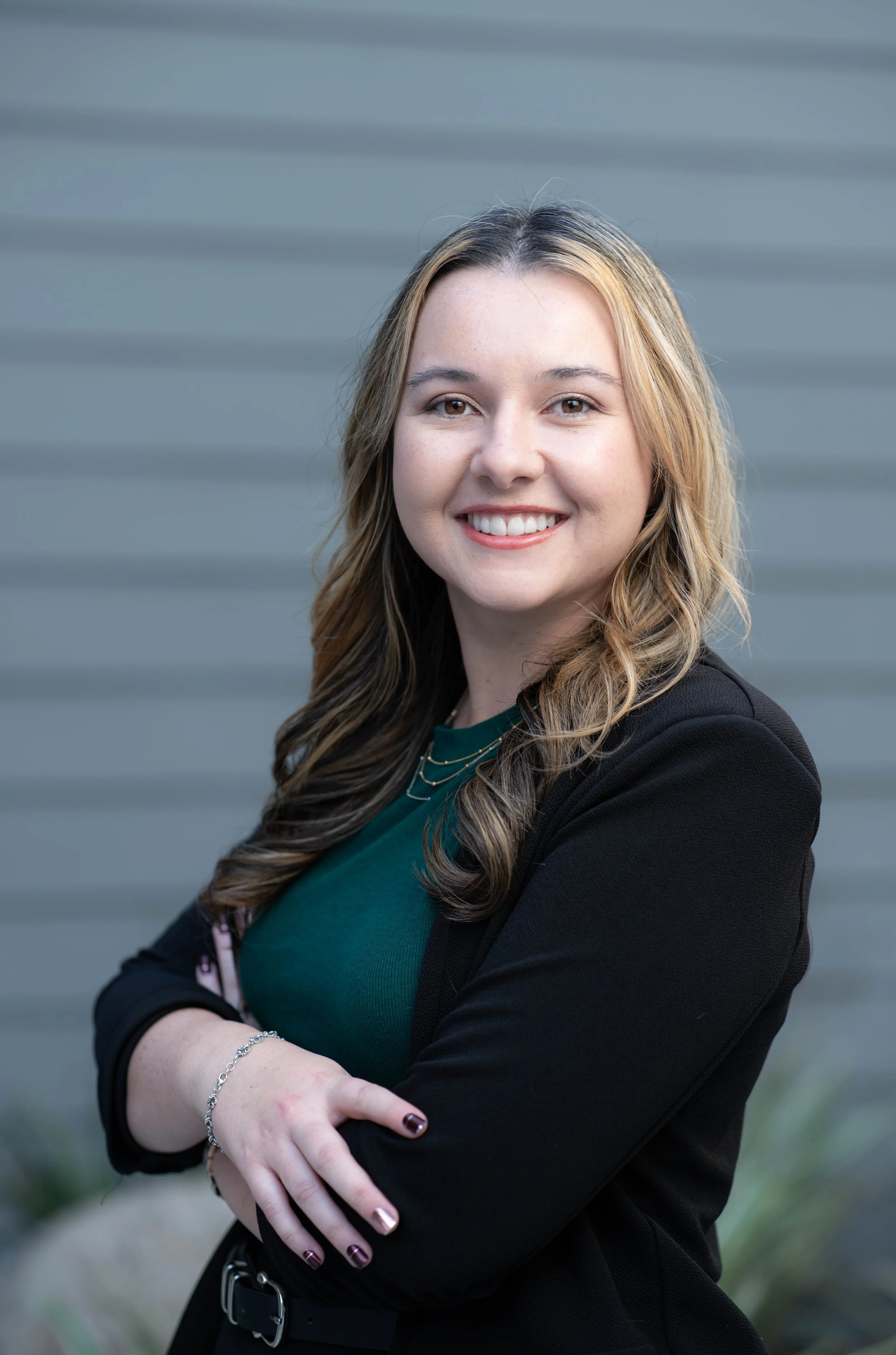 Portrait of a smiling woman with long wavy hair, wearing a green top, black blazer, and jewelry, standing in front of a gray, textured background.