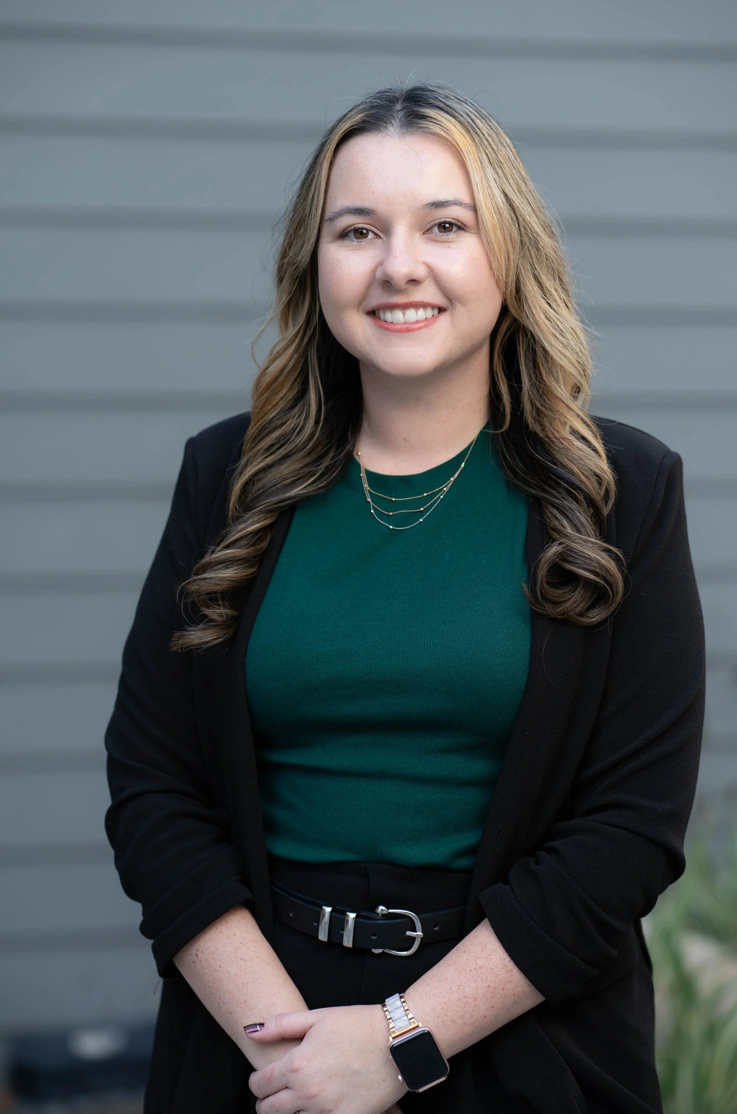 A woman with wavy brown hair wearing a black blazer, green top, and jewelry, standing in front of a gray building.