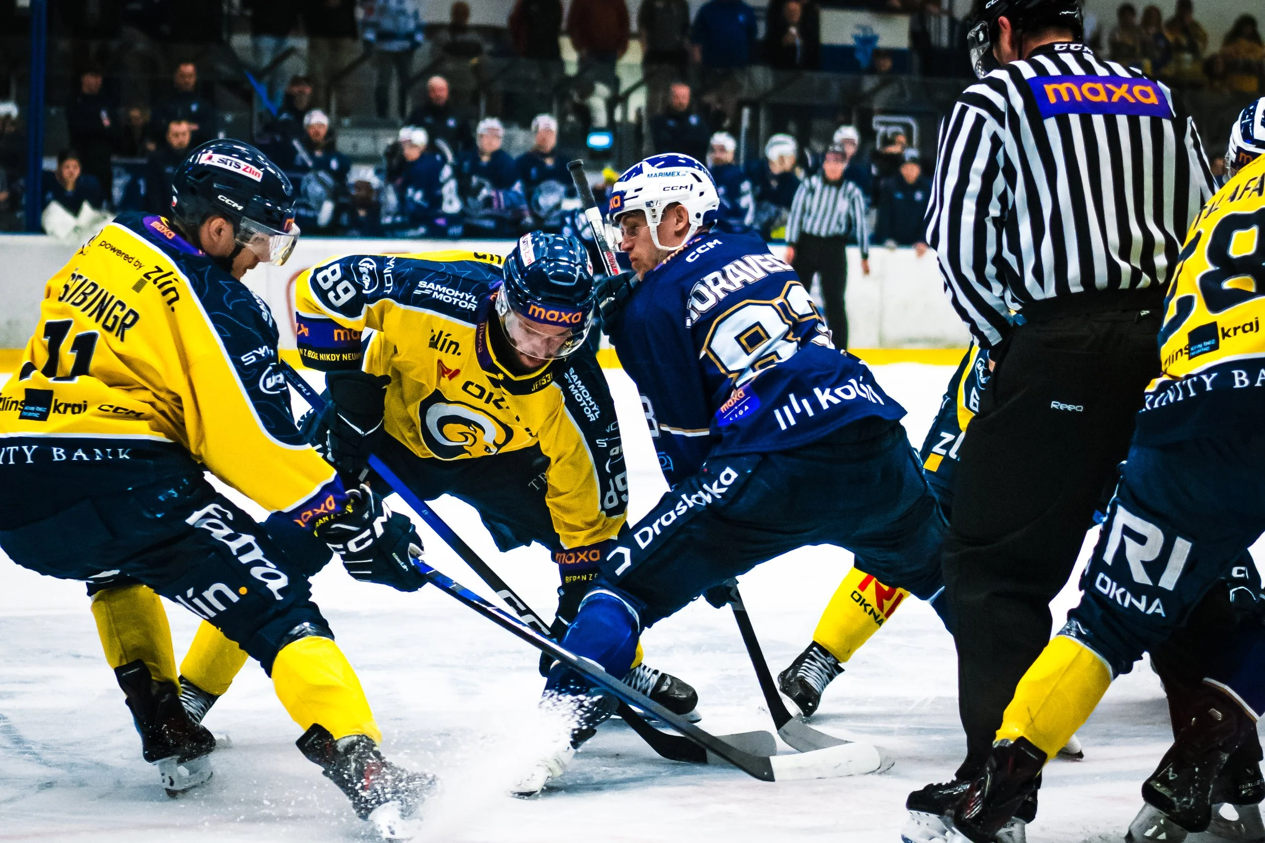 Hockey players from two teams battling for the puck during a game, with officials and spectators in the background.
