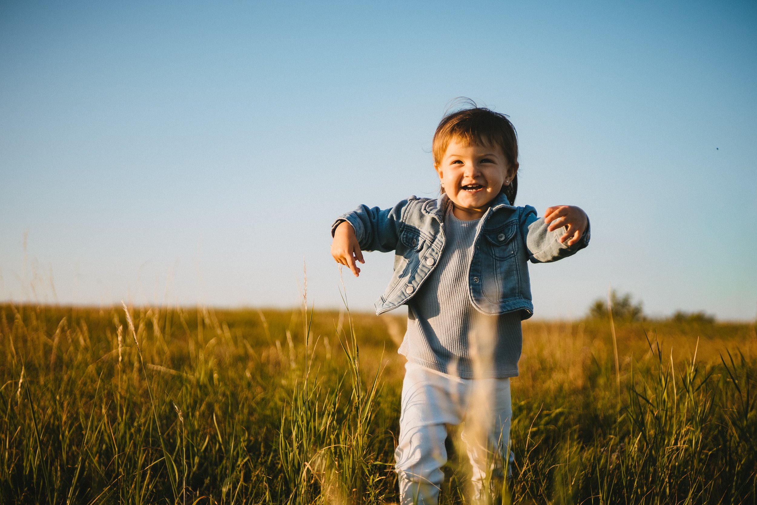 Child playing outdoors in a grassy field during sunset, smiling and reaching out.
