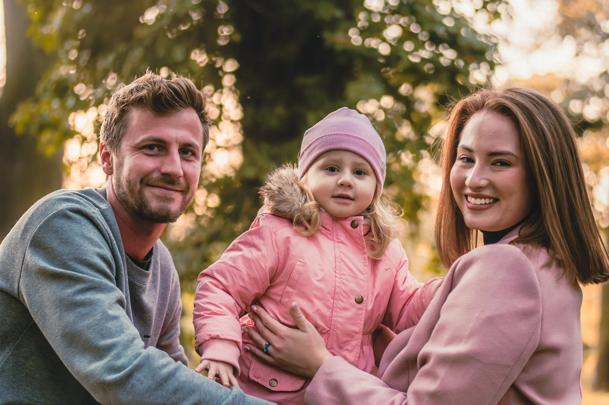 A family of three outdoors in a park during sunset, with a man, a woman, and a young girl dressed in pink jackets and warm clothing, smiling and looking at the camera.