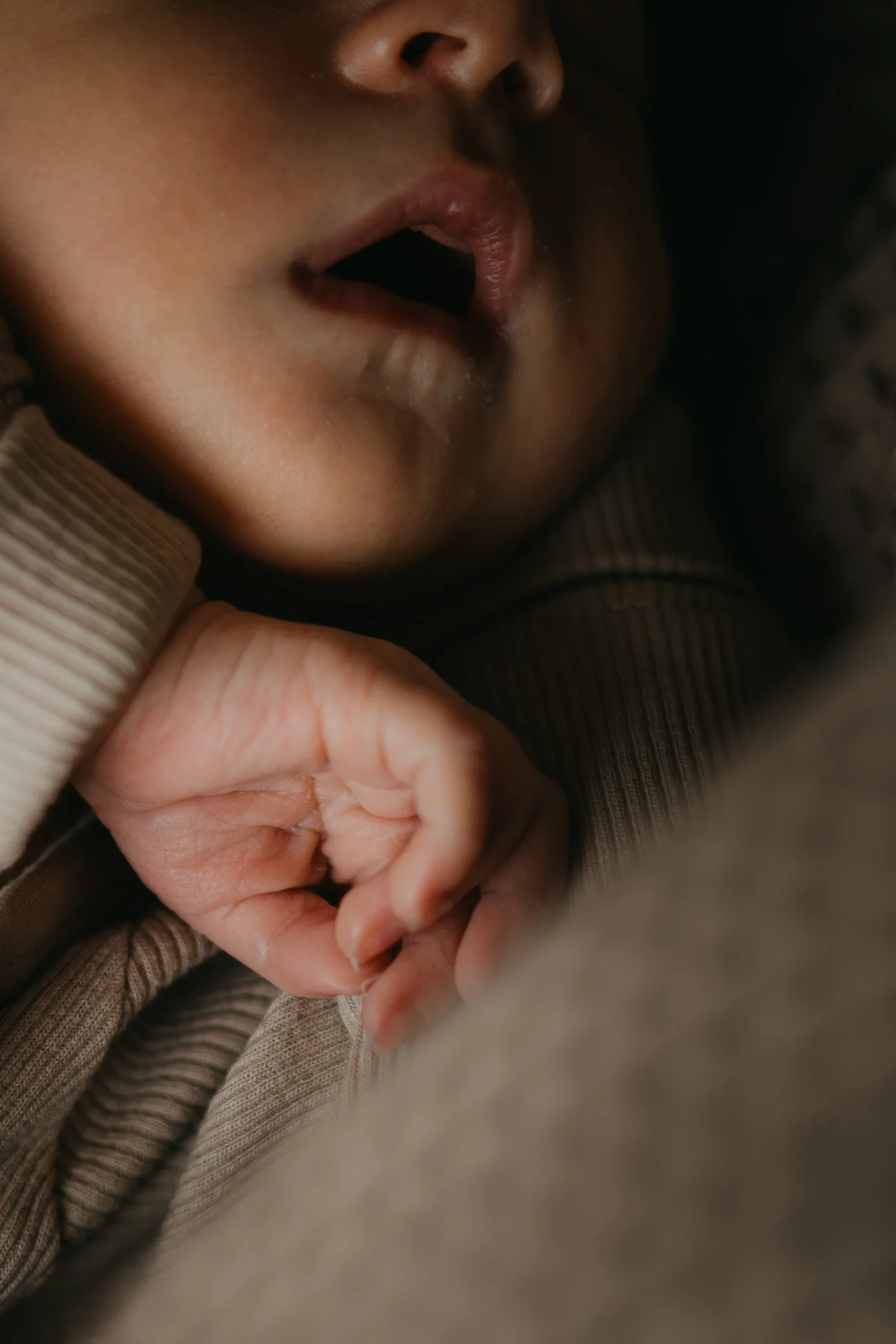 Close-up of a baby's face showing lips, part of the nose, and a hand resting near the chin, dressed in beige clothing.