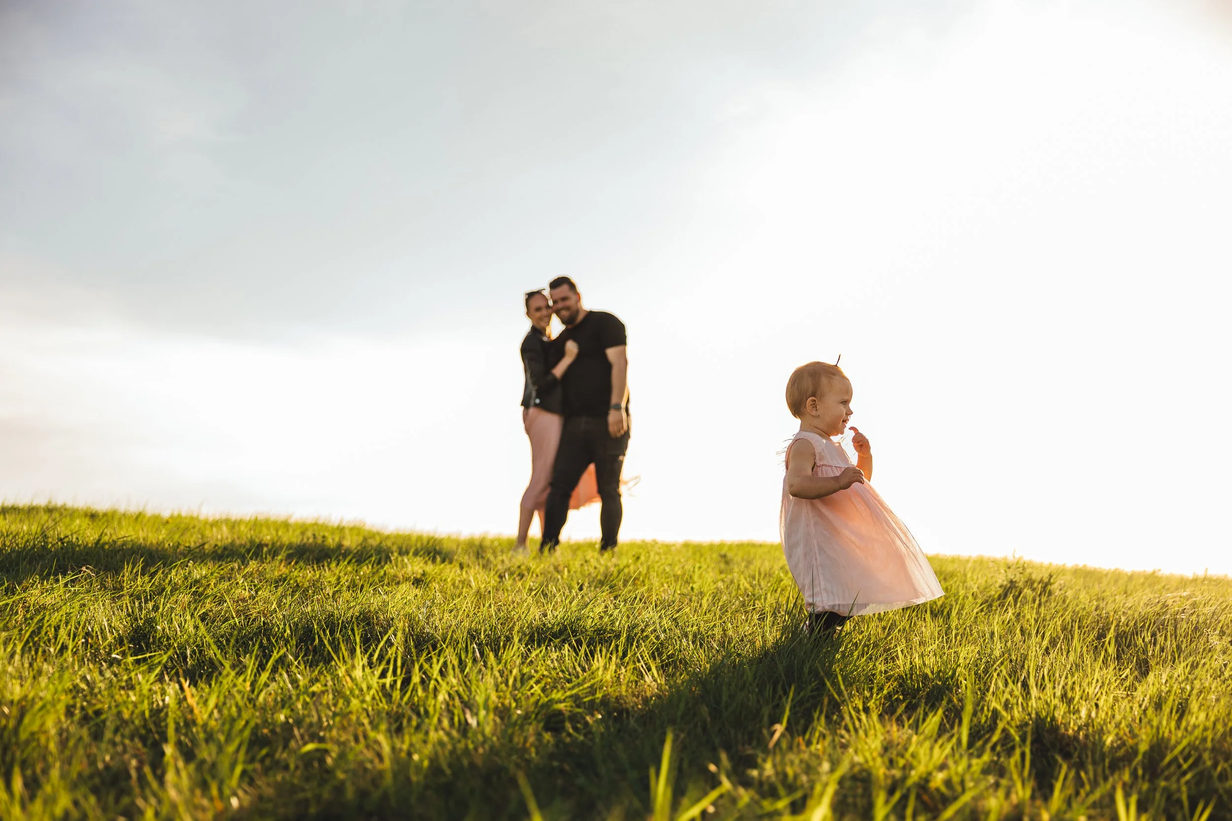 A young girl in a pink dress playing on a grassy hill with a couple in the background, under a bright sky during golden hour.
