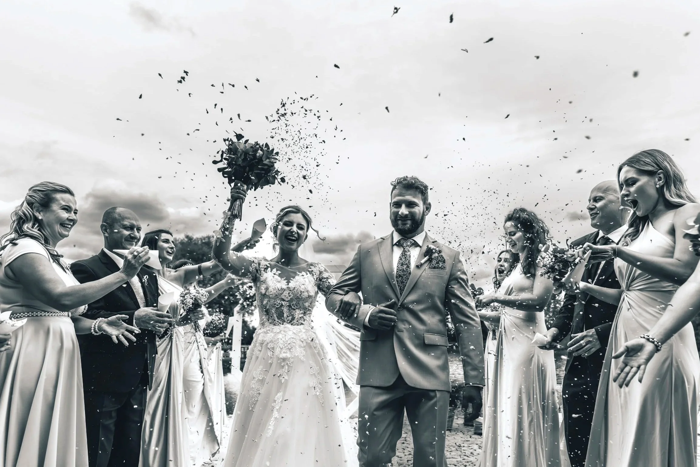 Black and white photo of a wedding celebration with bride and groom walking through guests throwing confetti, outdoors under cloudy sky.