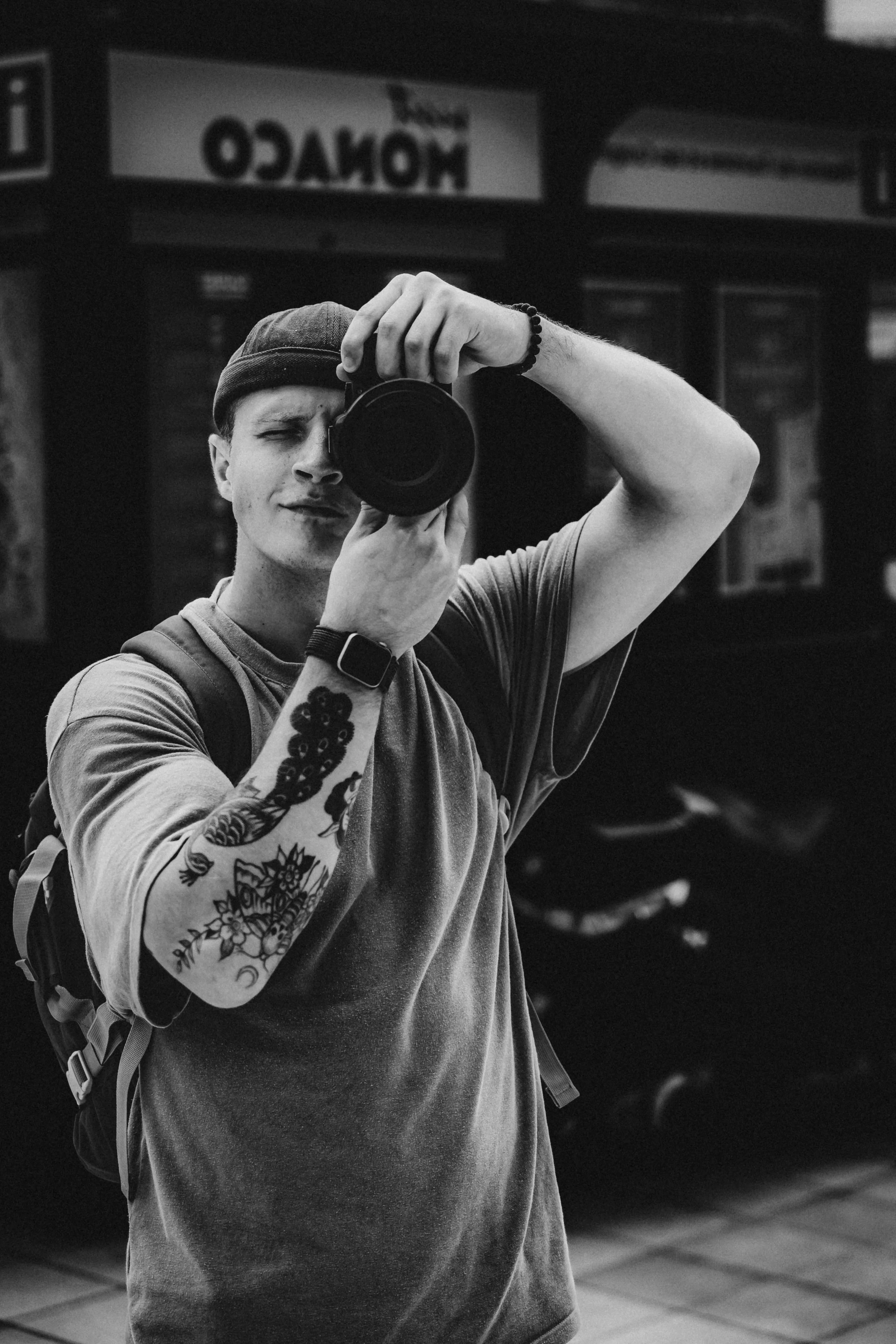 A young man with tattoos on his arm, wearing a beanie, taking a selfie in a mirror with a camera.