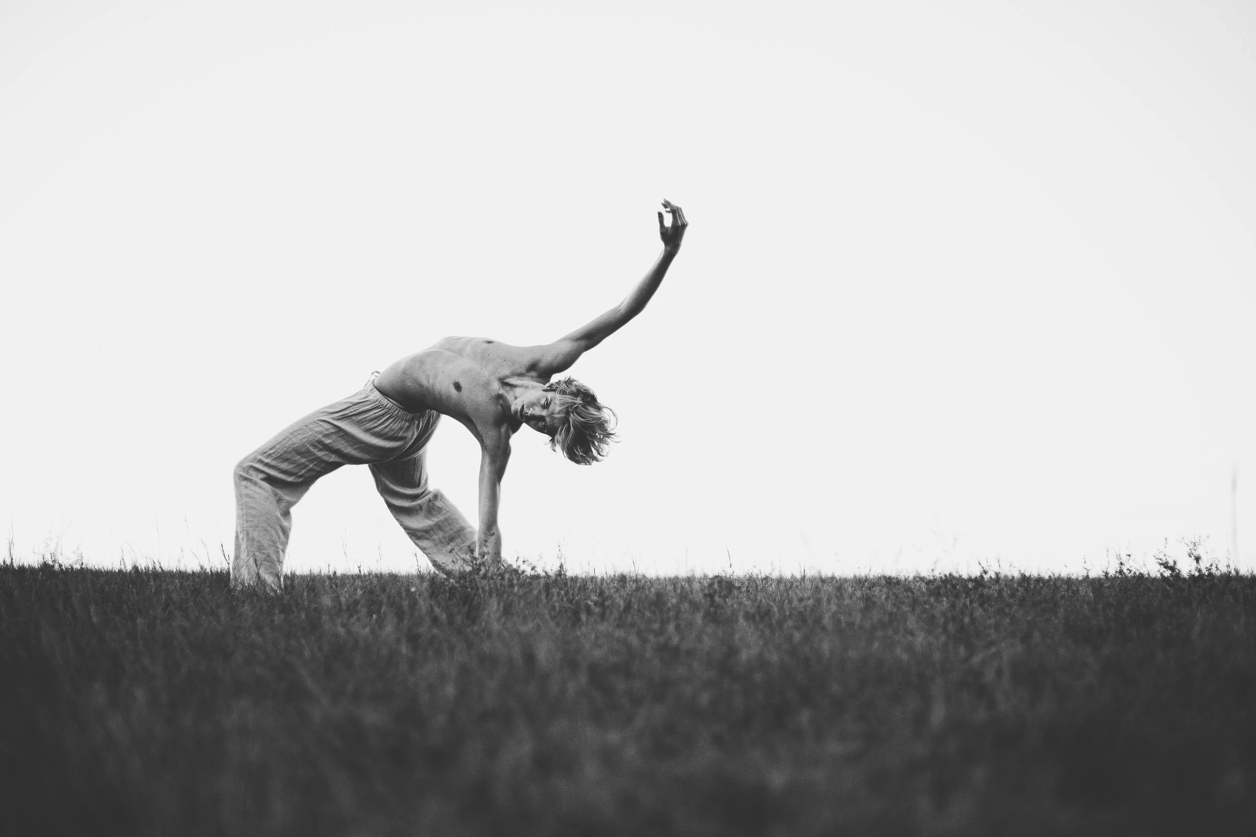 A shirtless man doing a yoga pose outdoors on grass, with one arm extended upward and the other hand on the ground, in black and white.