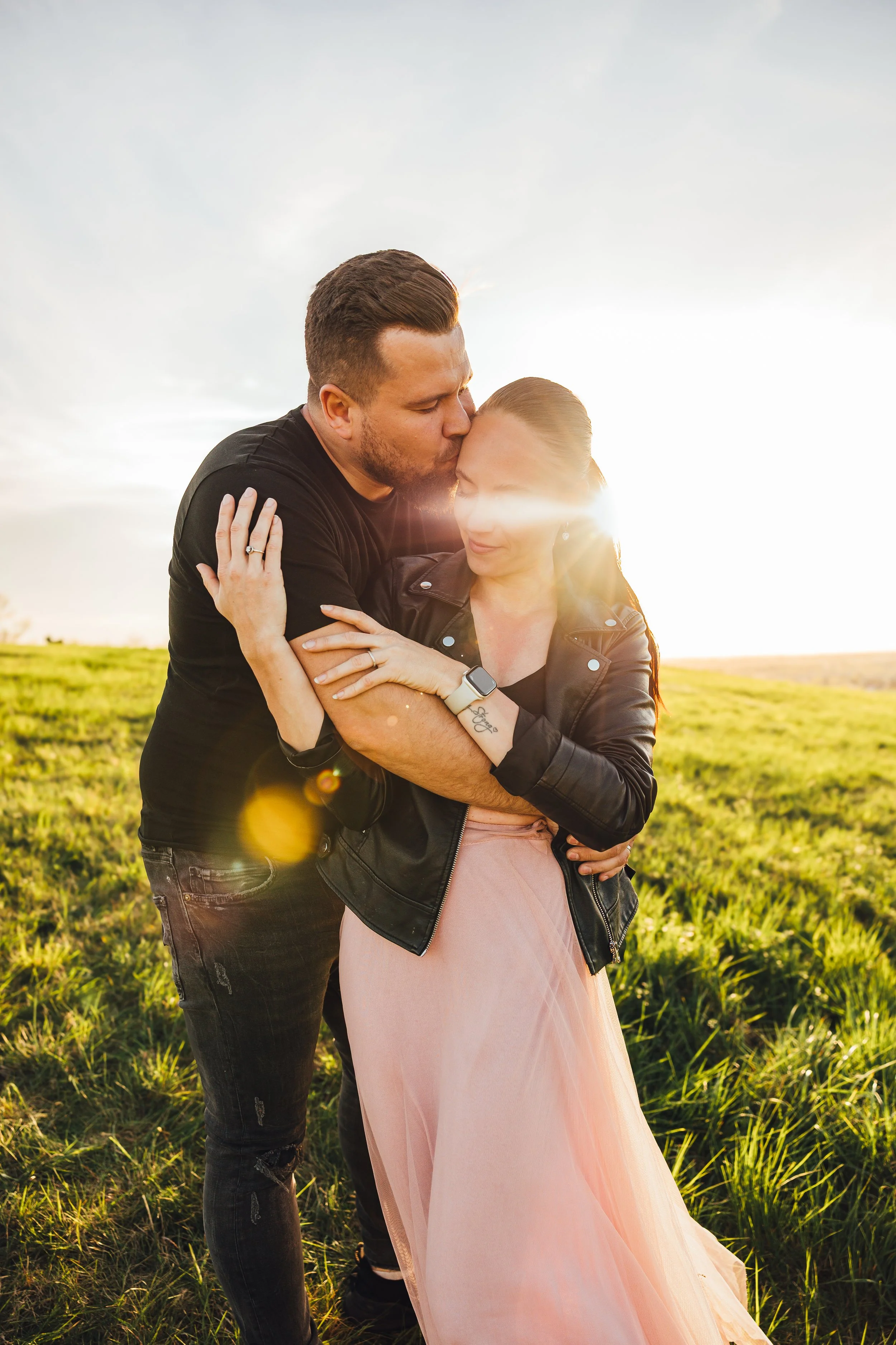 A couple embraces in a grassy field during sunset; the man is kissing the woman's forehead, both with eyes closed and content expressions.