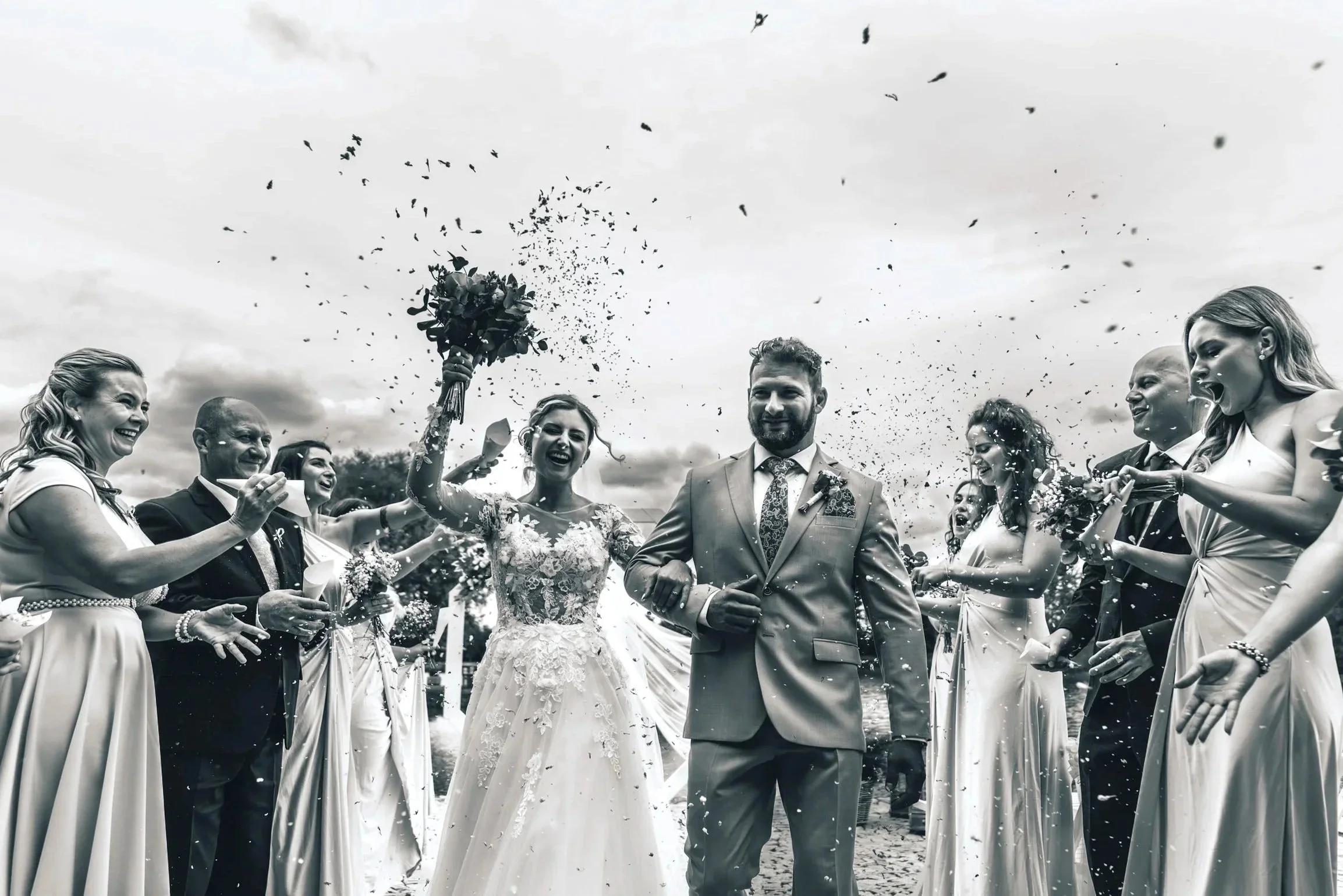 A bride and groom walking outdoors with friends and family celebrating around them, throwing confetti and smiling.