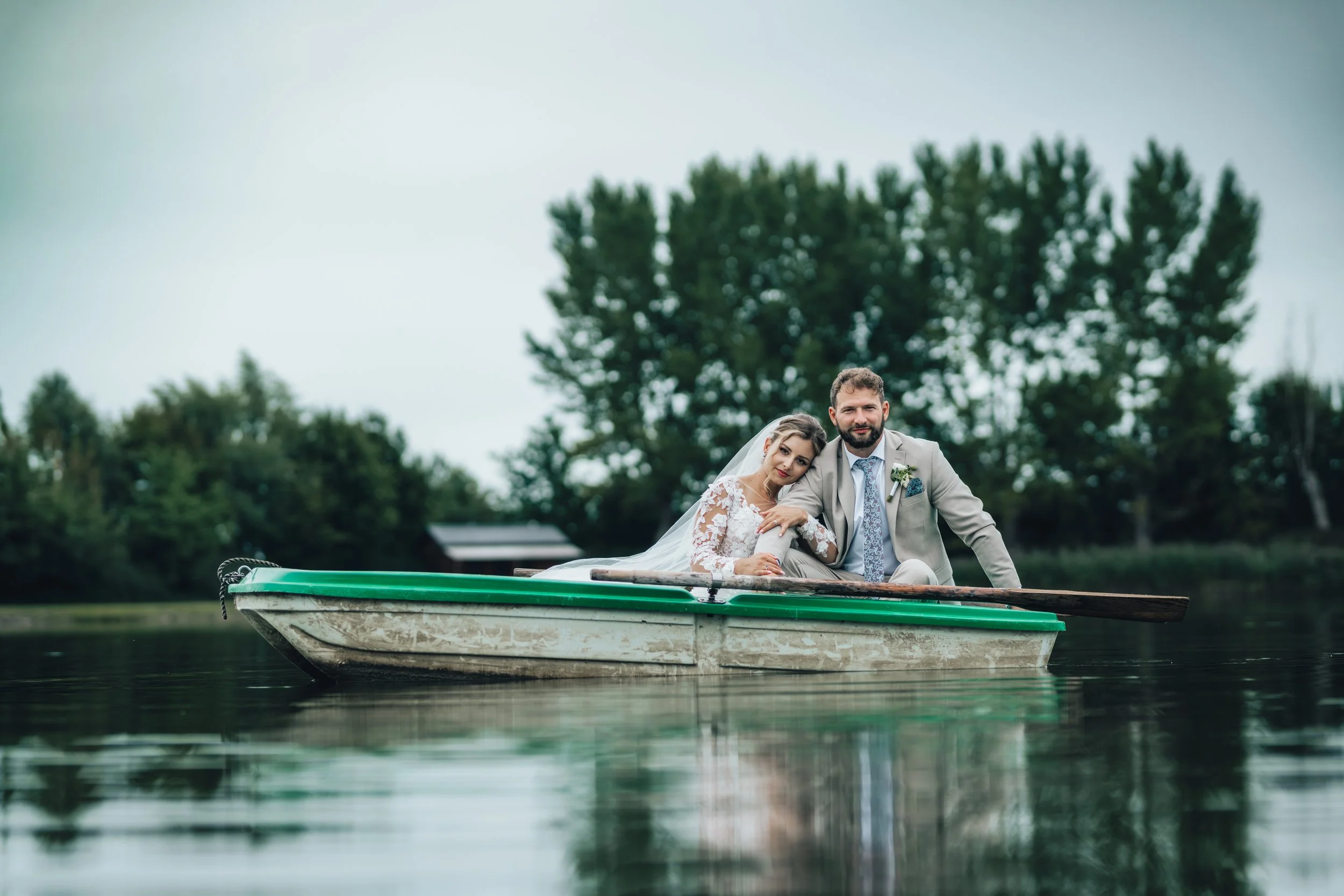 Bride and groom sitting in a rowboat on a calm lake, surrounded by trees, with the bride resting her head on the groom's shoulder.