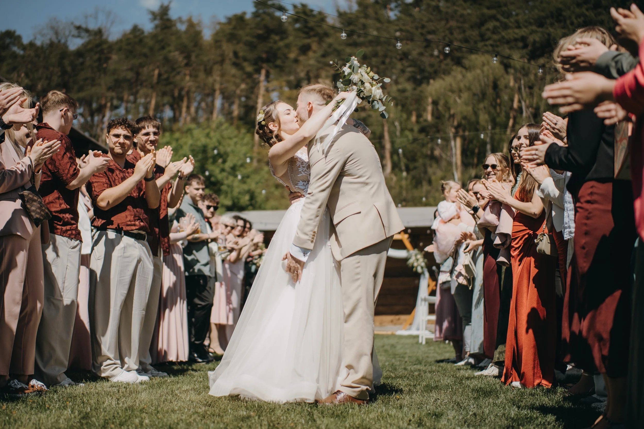 A bride and groom kiss at an outdoor wedding ceremony, surrounded by wedding guests clapping and smiling, with trees and string lights in the background.