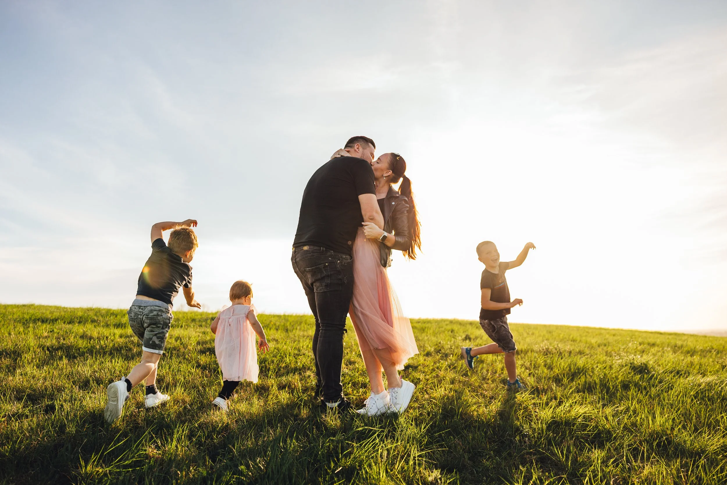 A family of five, including two children and two adults, sharing a kiss on a grassy field during sunset, with three children playing and running nearby.