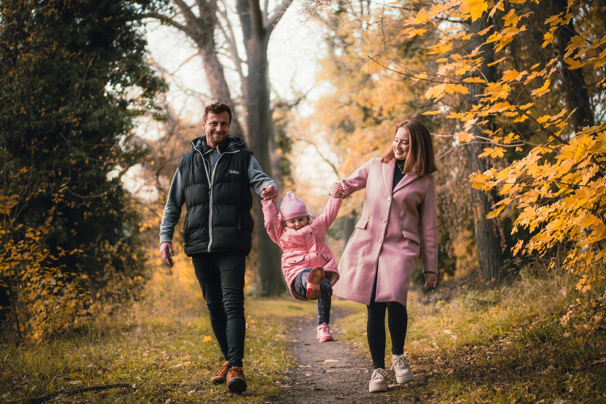 A family of three, consisting of a man, a woman, and a young girl, walking along a fall foliage trail. The man and woman are holding the child's hands as she swings between them. The scene features autumn leaves and trees with yellow and orange folia