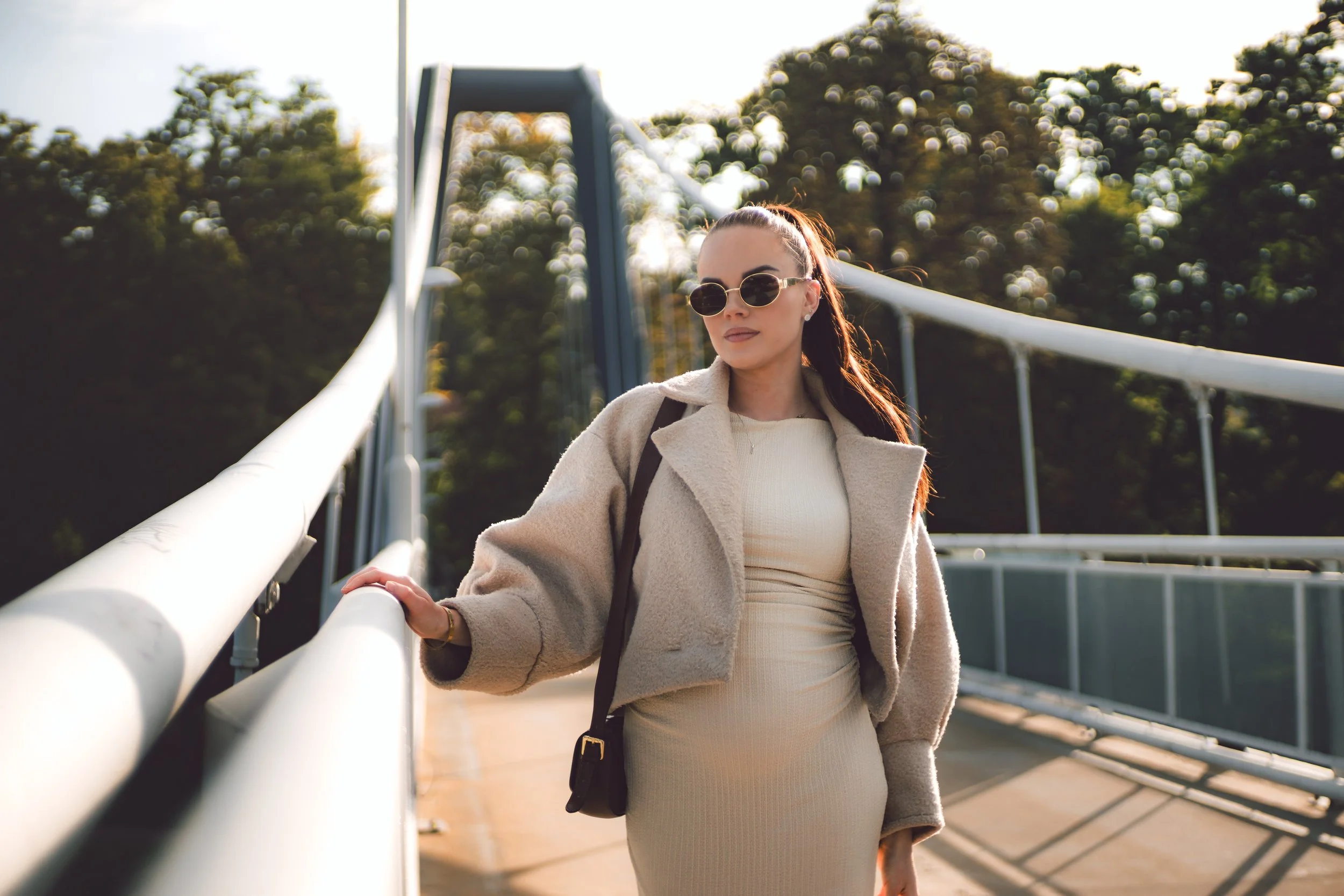 A woman wearing sunglasses, a beige dress, and a beige coat, standing on a bridge with her hand on the railing, with trees and blue sky in the background.