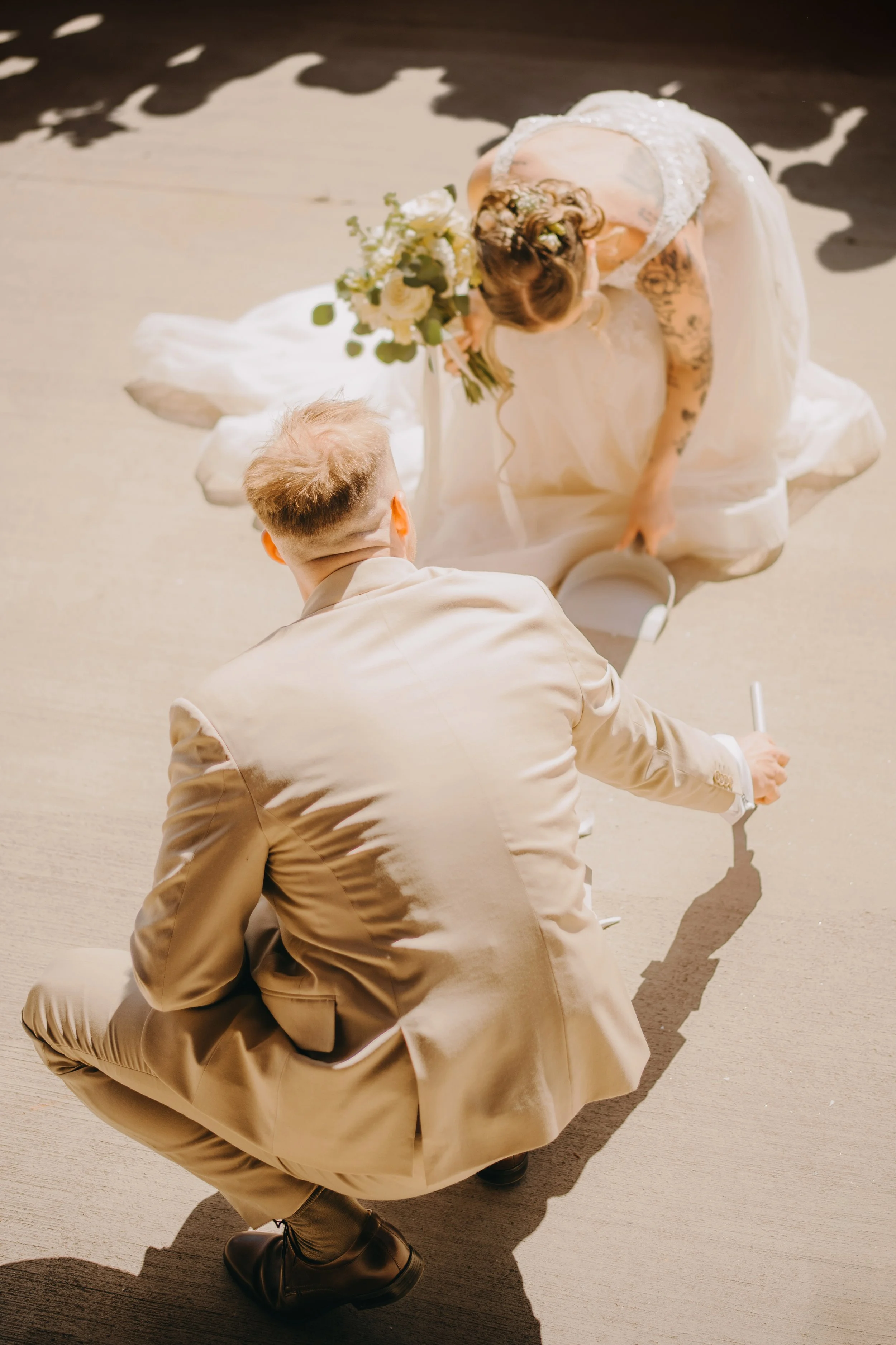 A bride and groom, dressed in white and beige, are kneeling on the floor, with the bride bowing her head and holding a bouquet of white flowers. The groom is facing away, in a beige suit, with his head bowed, on a plain beige floor.