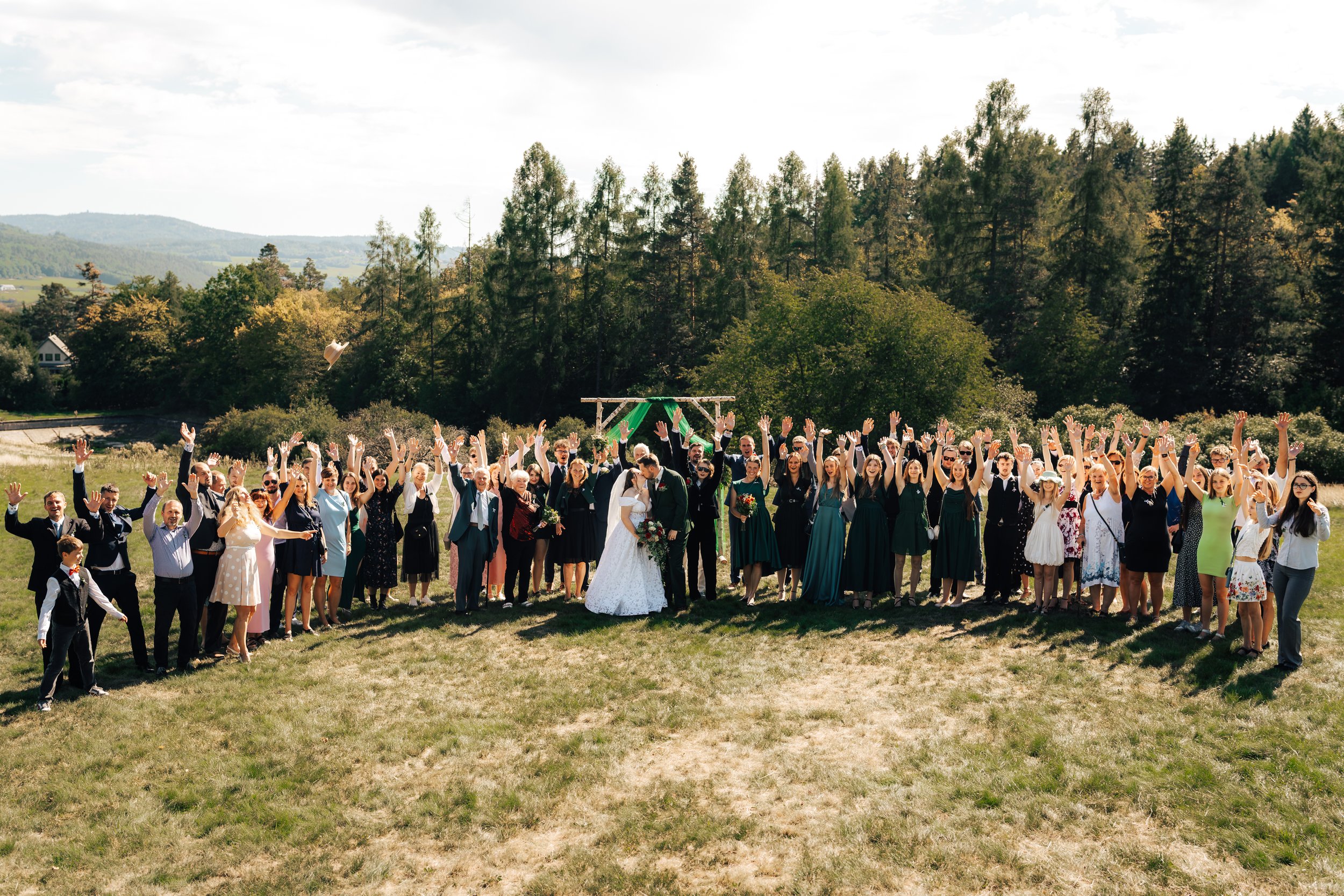 A large group of wedding guests outdoors in a grassy field with mountains and trees in the background, celebrating a wedding with raised hands and a bride and groom in the center.