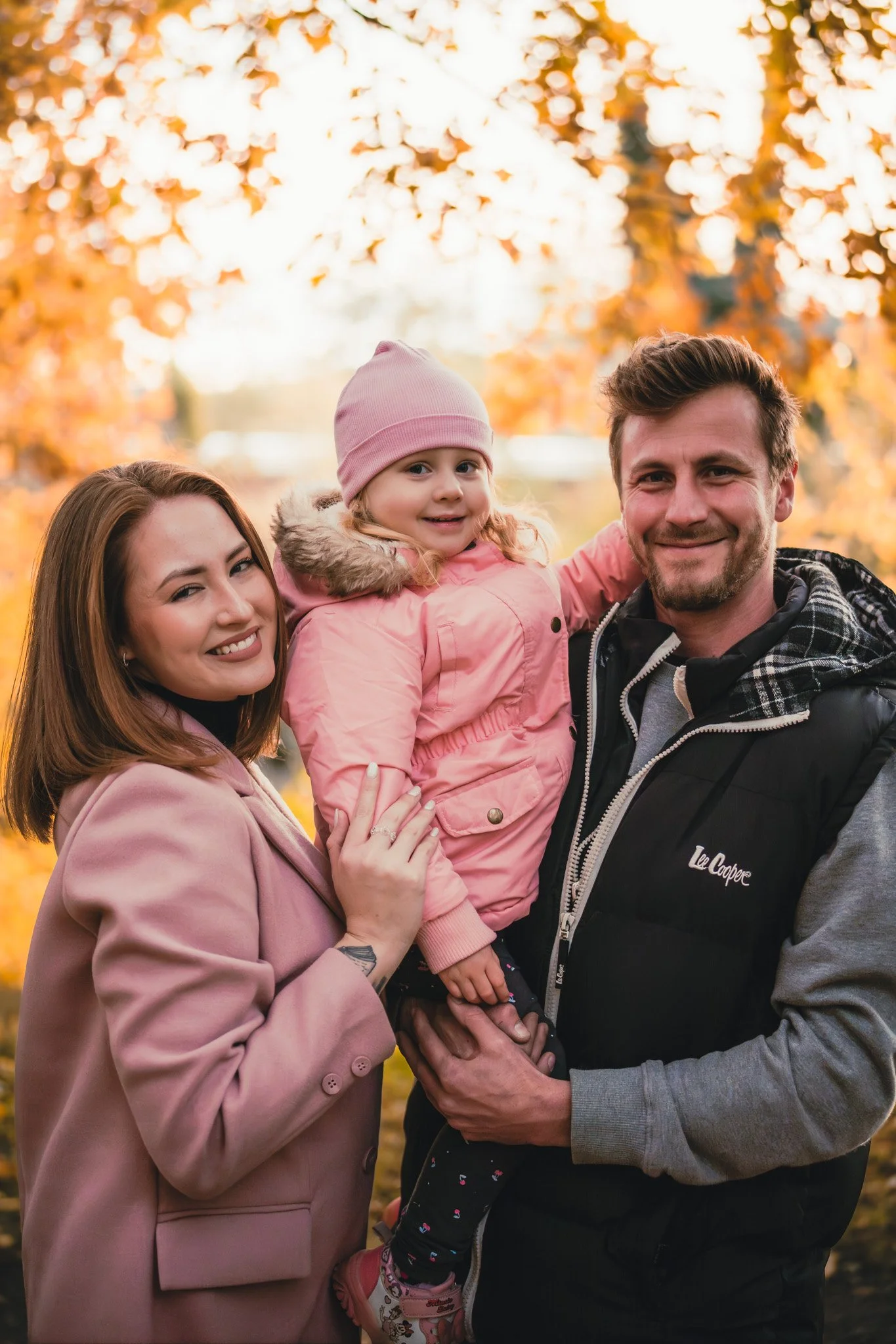 A family of three smiling outdoors during fall with colorful autumn leaves in the background. The mother has brown hair and is wearing a pink coat, the father has brown hair and is wearing a black vest over a gray hoodie, and their young daughter, in
