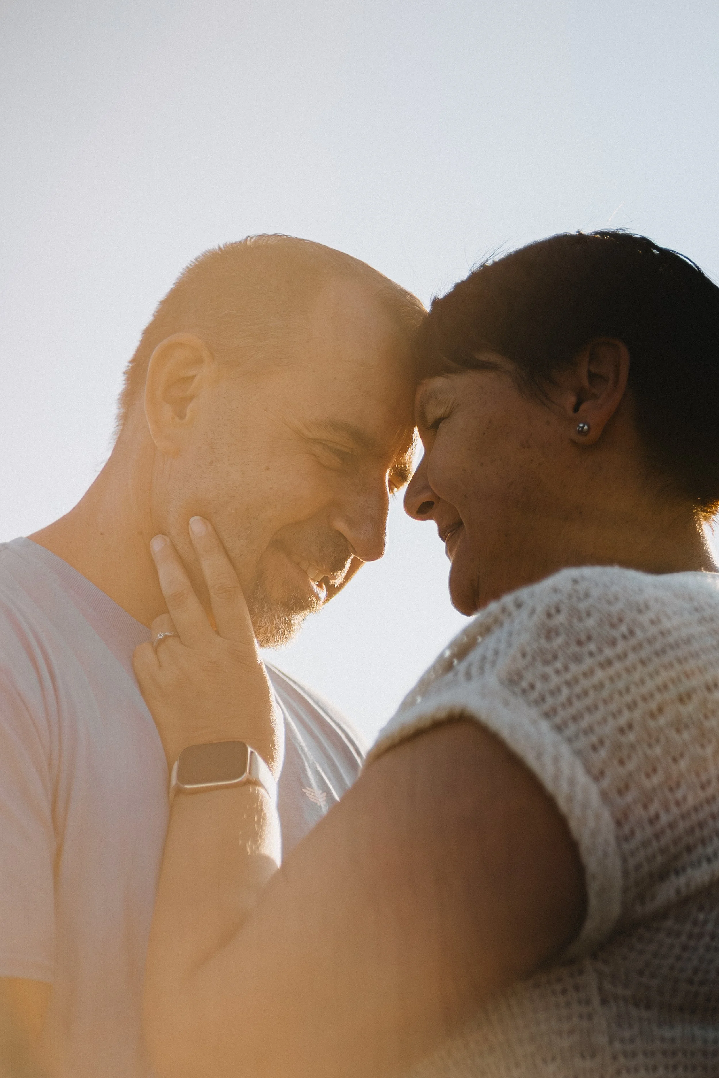 A couple shares a tender moment with foreheads touching and eyes closed, enjoying each other's presence outdoors with sunlight in the background.