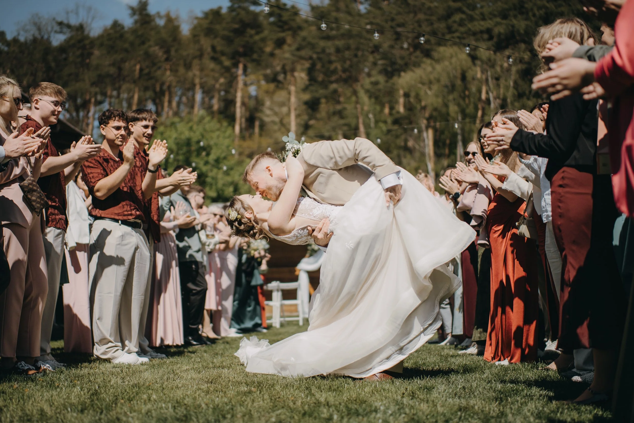 A newlywed couple sharing a dance during their outdoor wedding reception, surrounded by guests clapping and smiling under string lights in a green natural setting.