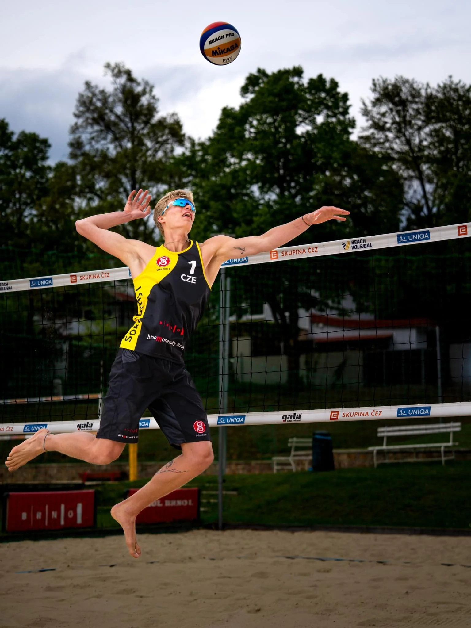 A woman in a black and yellow sports uniform playing beach volleyball jumps to hit a volleyball in mid-air, with trees and a volleyball court in the background.