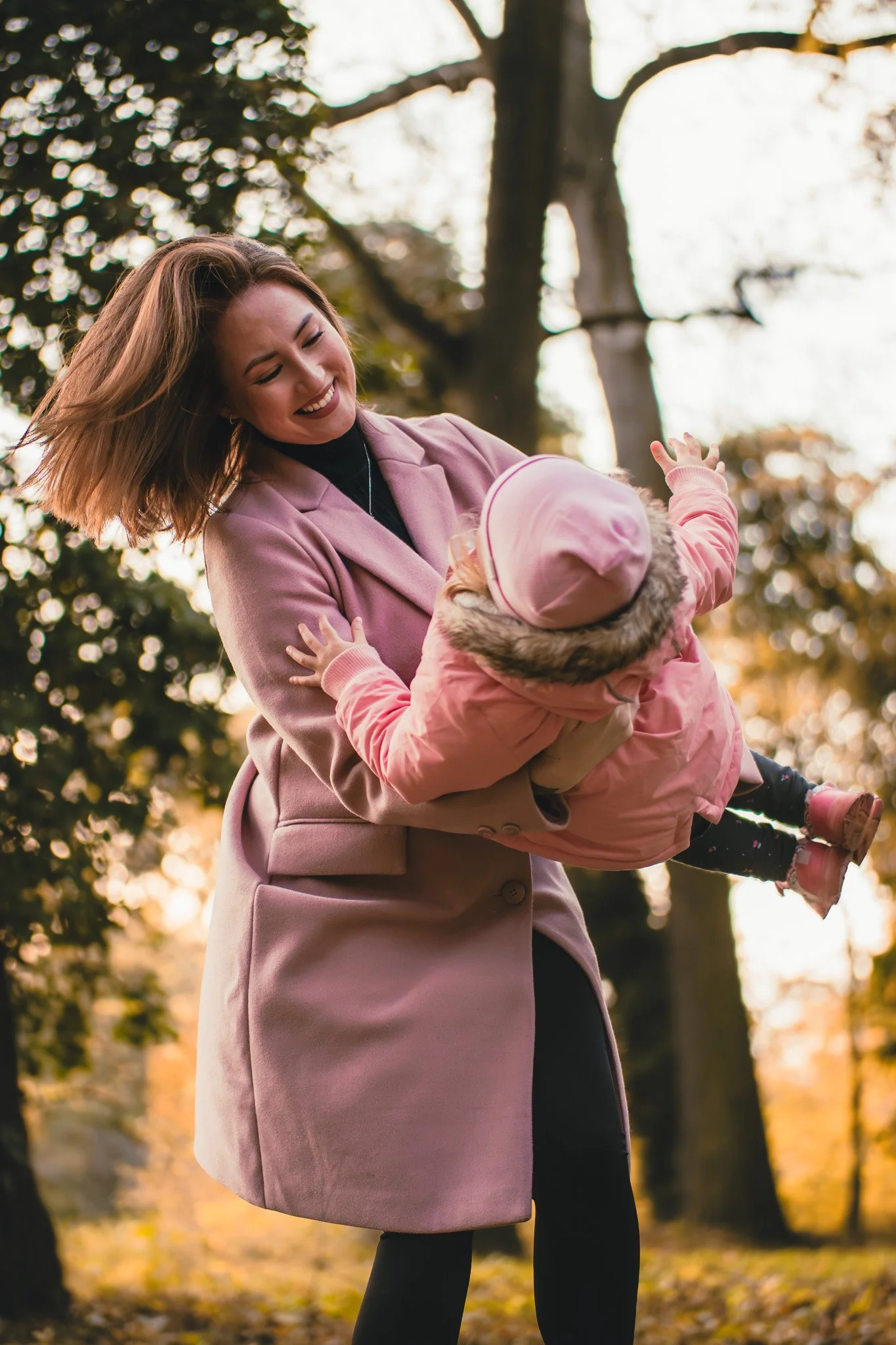 A woman wearing a pink coat happily playing with a young child dressed in pink outdoors during autumn.