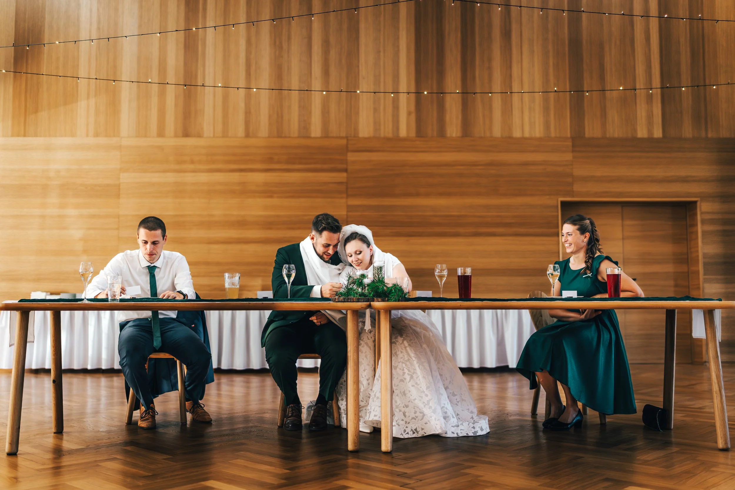 Wedding reception scene with three people seated at a long table. The bride and groom are in the middle, sharing a moment looking at something together, while a woman sits on the right and a man on the left. The table is decorated with a small greene