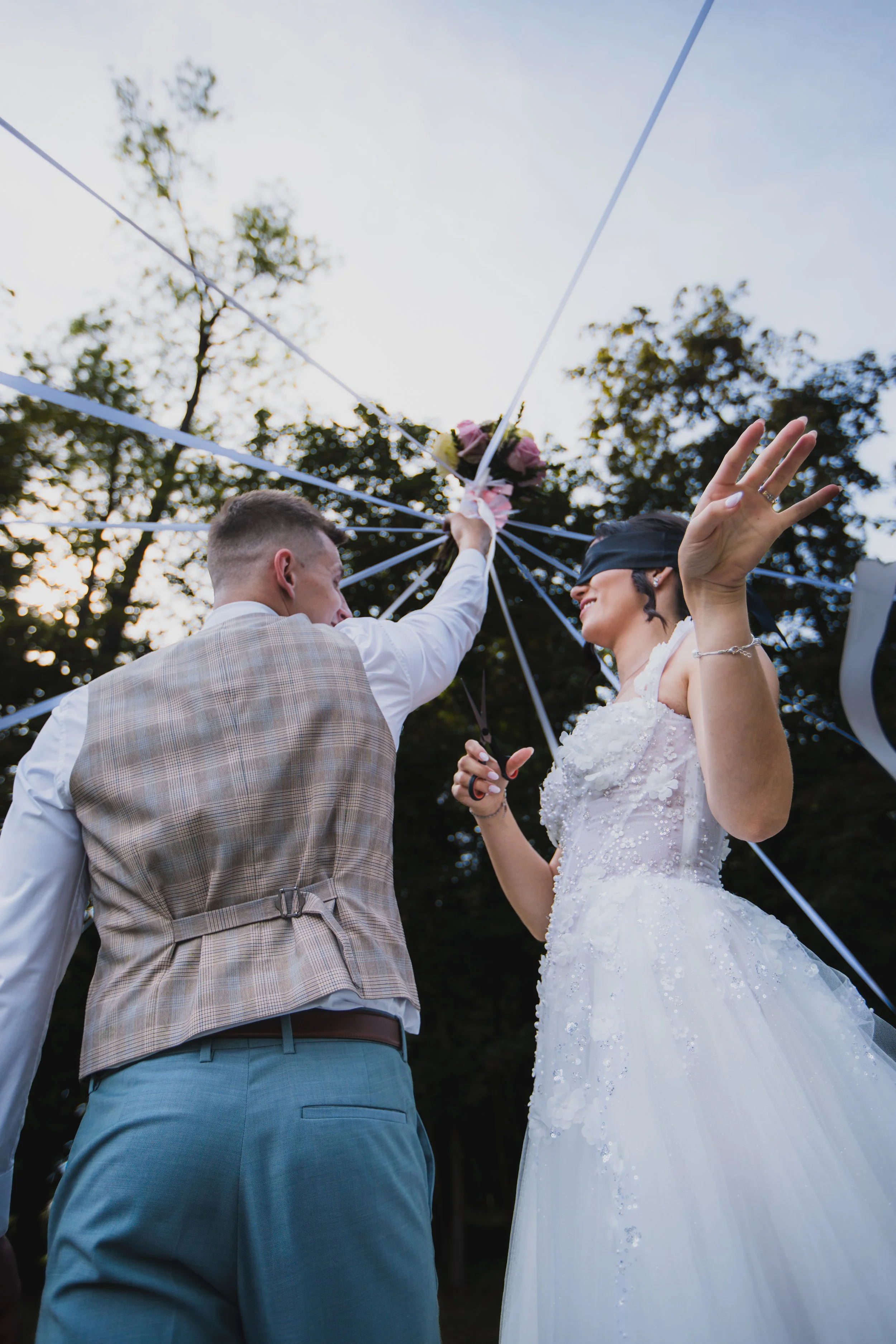A bride and groom during their wedding ceremony, with the groom placing a bouquet on the bride's head while both are smiling; the bride is wearing a white wedding dress and a blindfold, and the groom is in a vest and shirt.