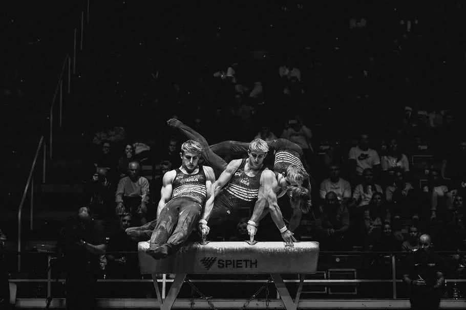 Three male gymnasts performing a synchronized routine on a pommel horse during a gymnastics competition, with an audience watching in the background.