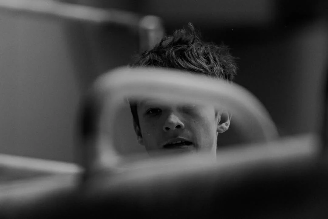 A young boy with messy hair looking into a mirror with a blurred foreground.