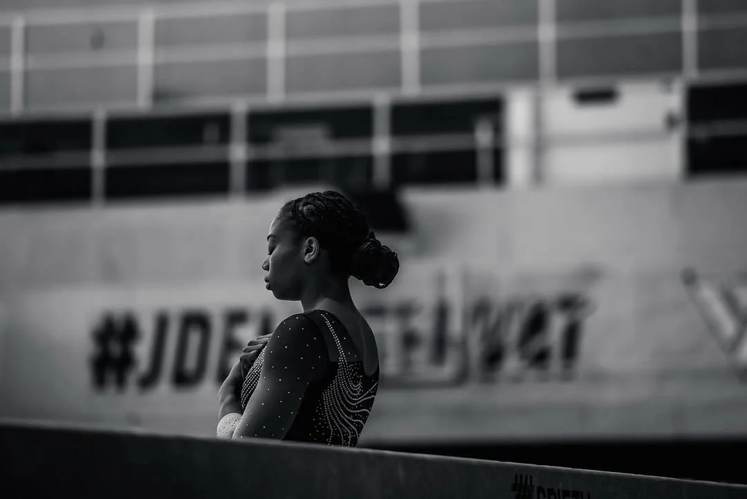 A gymnast in a glittery leotard standing with arms crossed, looking downward, in an indoor sports arena.
