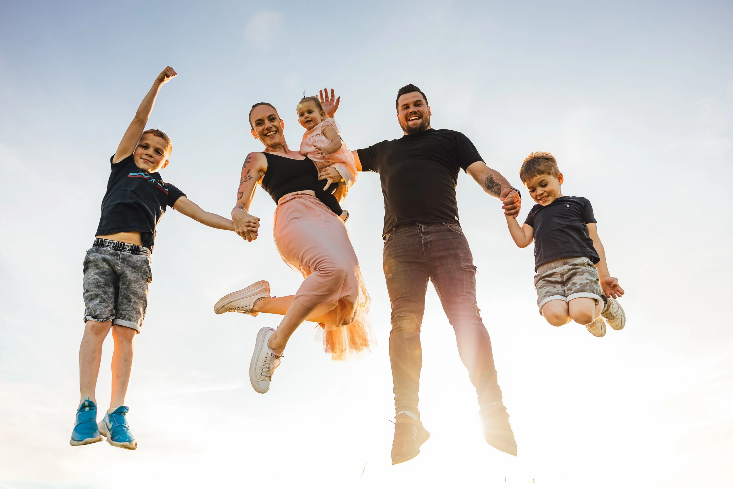 A family of six holding hands and jumping in the air outdoors during sunset, smiling and enjoying time together.