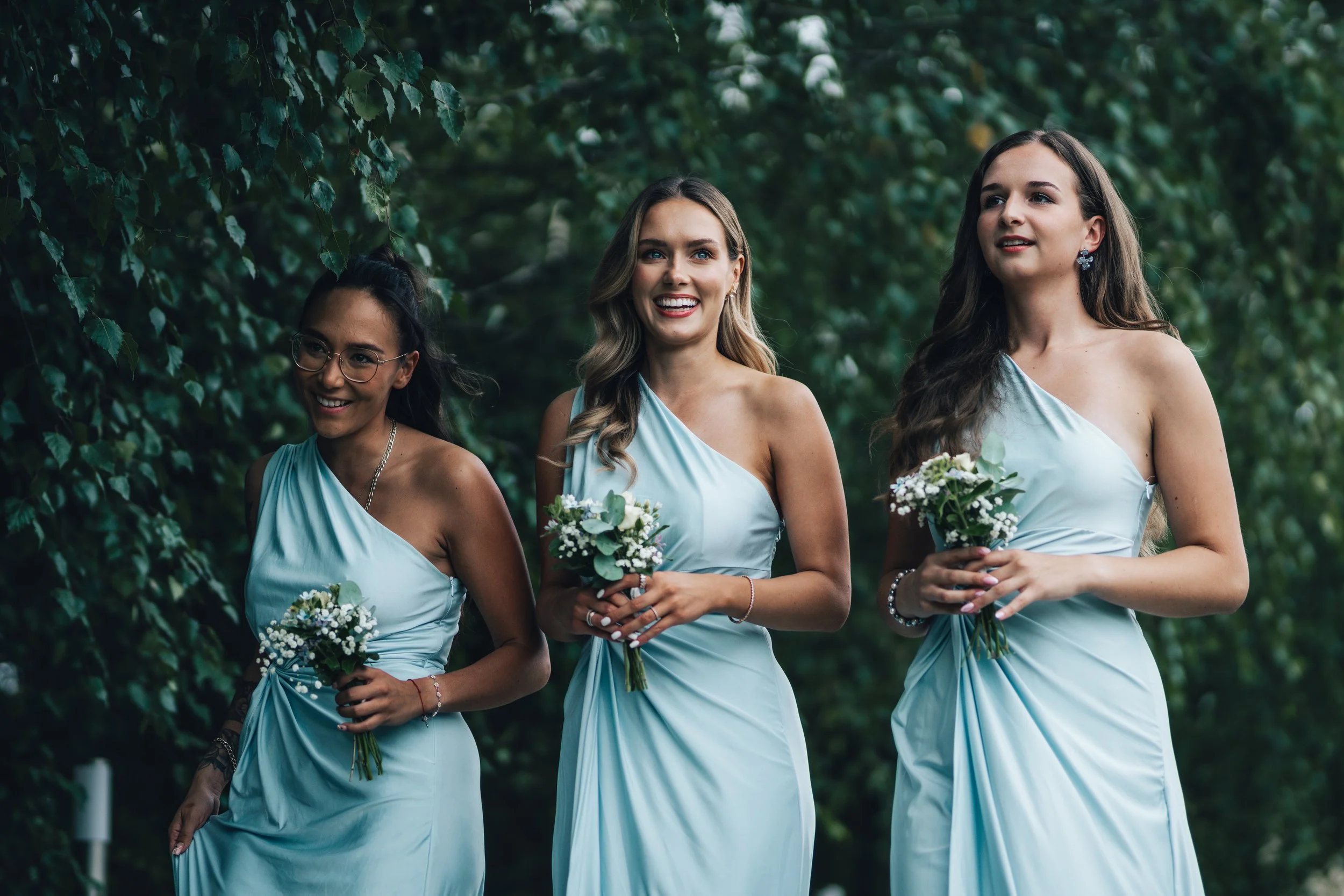 Three women in light blue dresses holding small bouquets of white flowers outdoors surrounded by green trees.