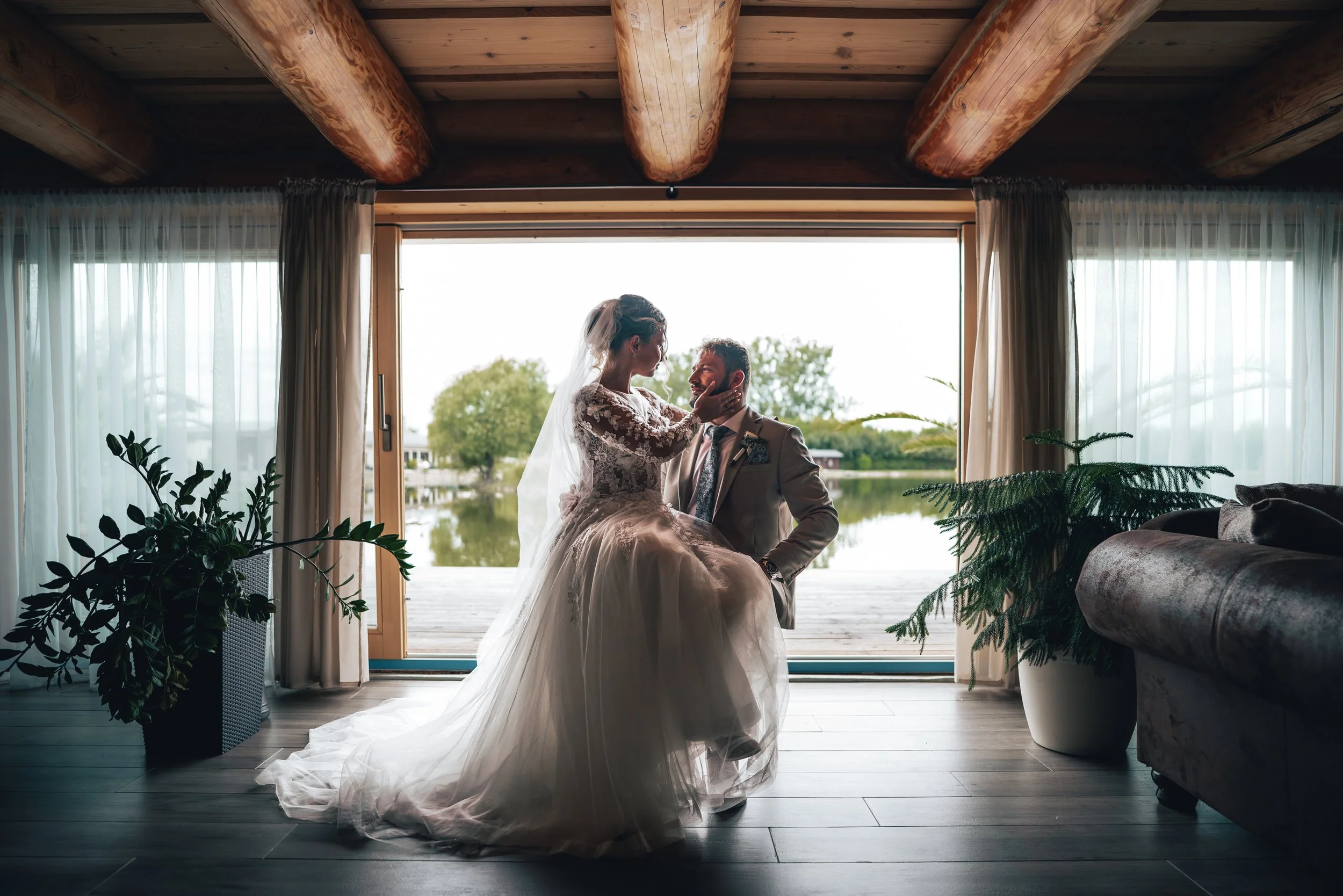 A bride and groom in wedding attire inside a room with large windows overlooking a lake and trees, with plants and furniture nearby.
