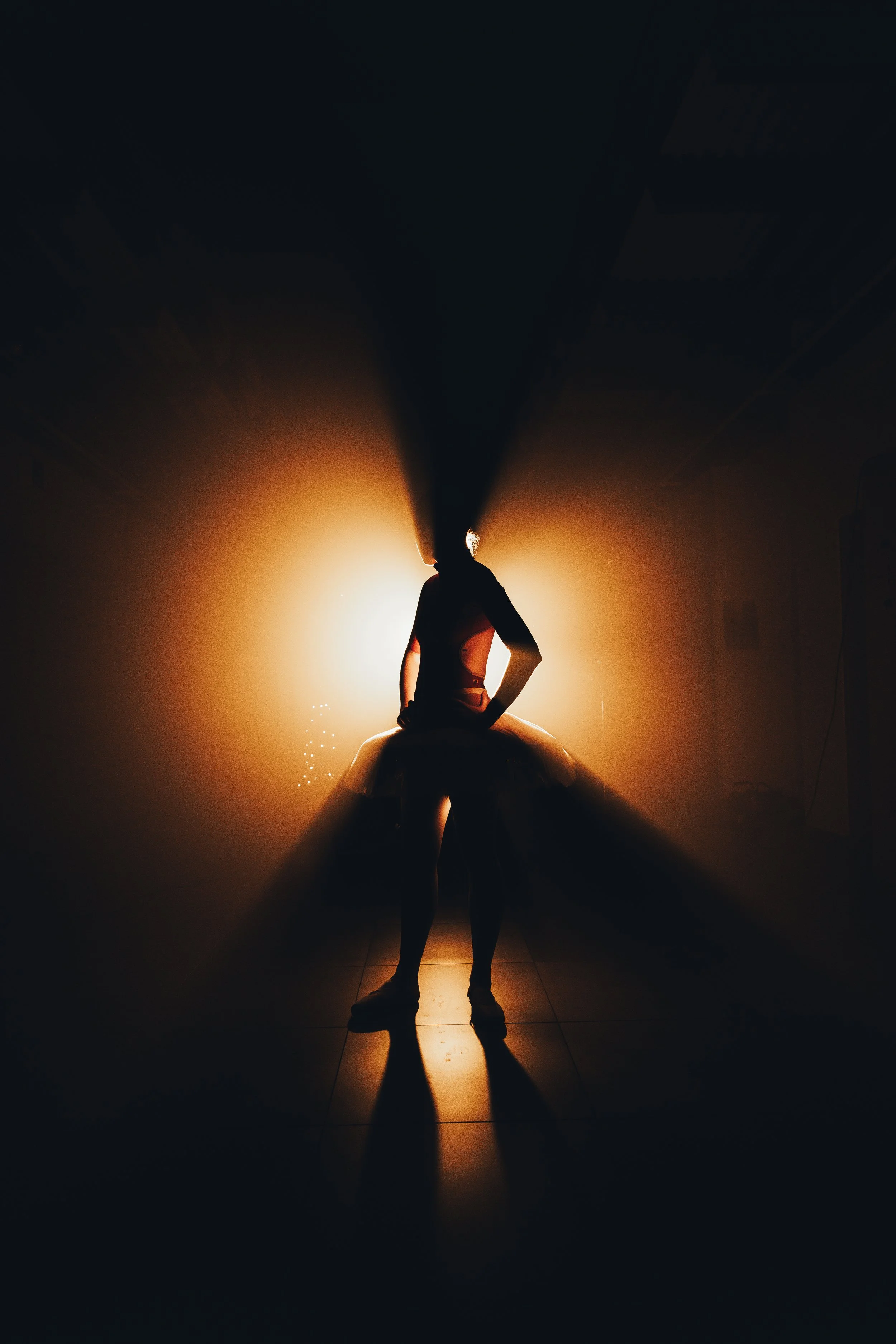 Silhouette of a girl in a tutu standing in front of a bright backlight, creating long shadows on the floor.