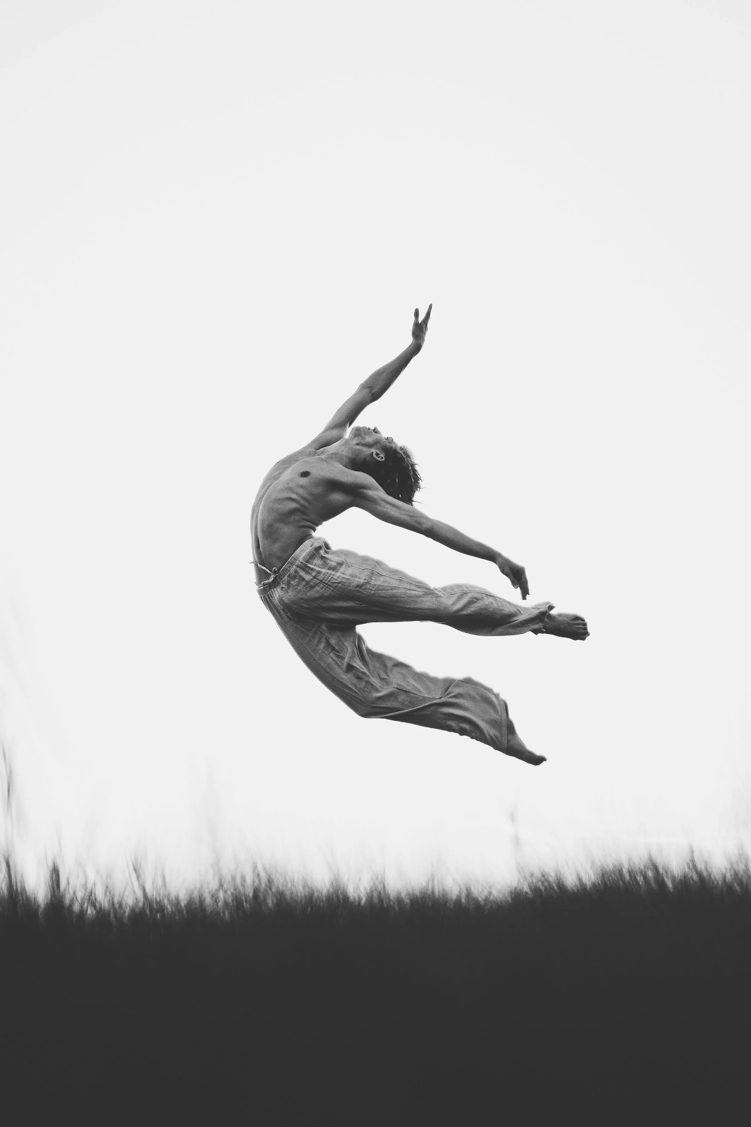 A shirtless person jumps high in the air outdoors, with arms and legs extended, against a plain sky above a grassy field.