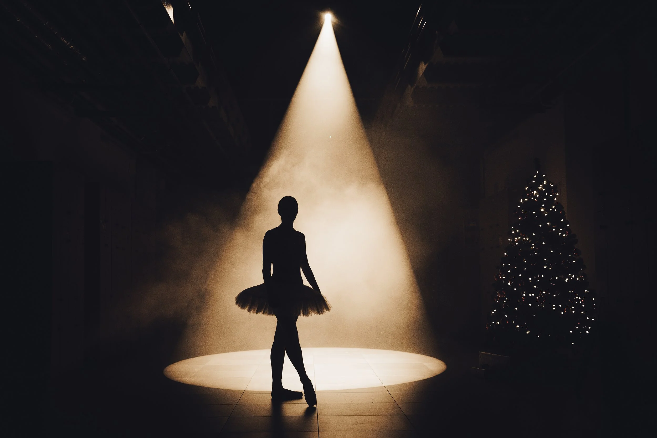 Ballet dancer silhouette standing on stage with spotlight, Christmas tree with lights in the background.