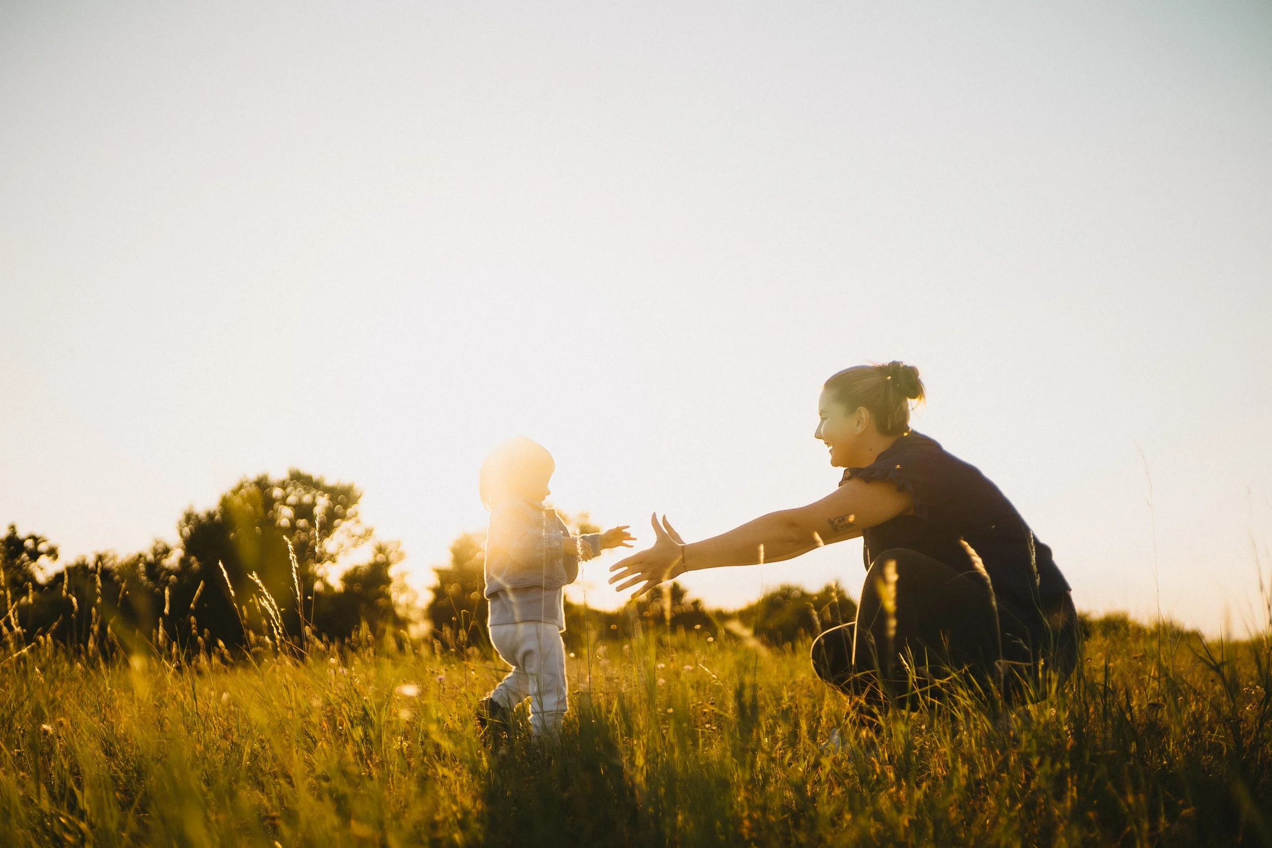 A woman and a small child playing and reaching out to each other in a grassy field during sunset.