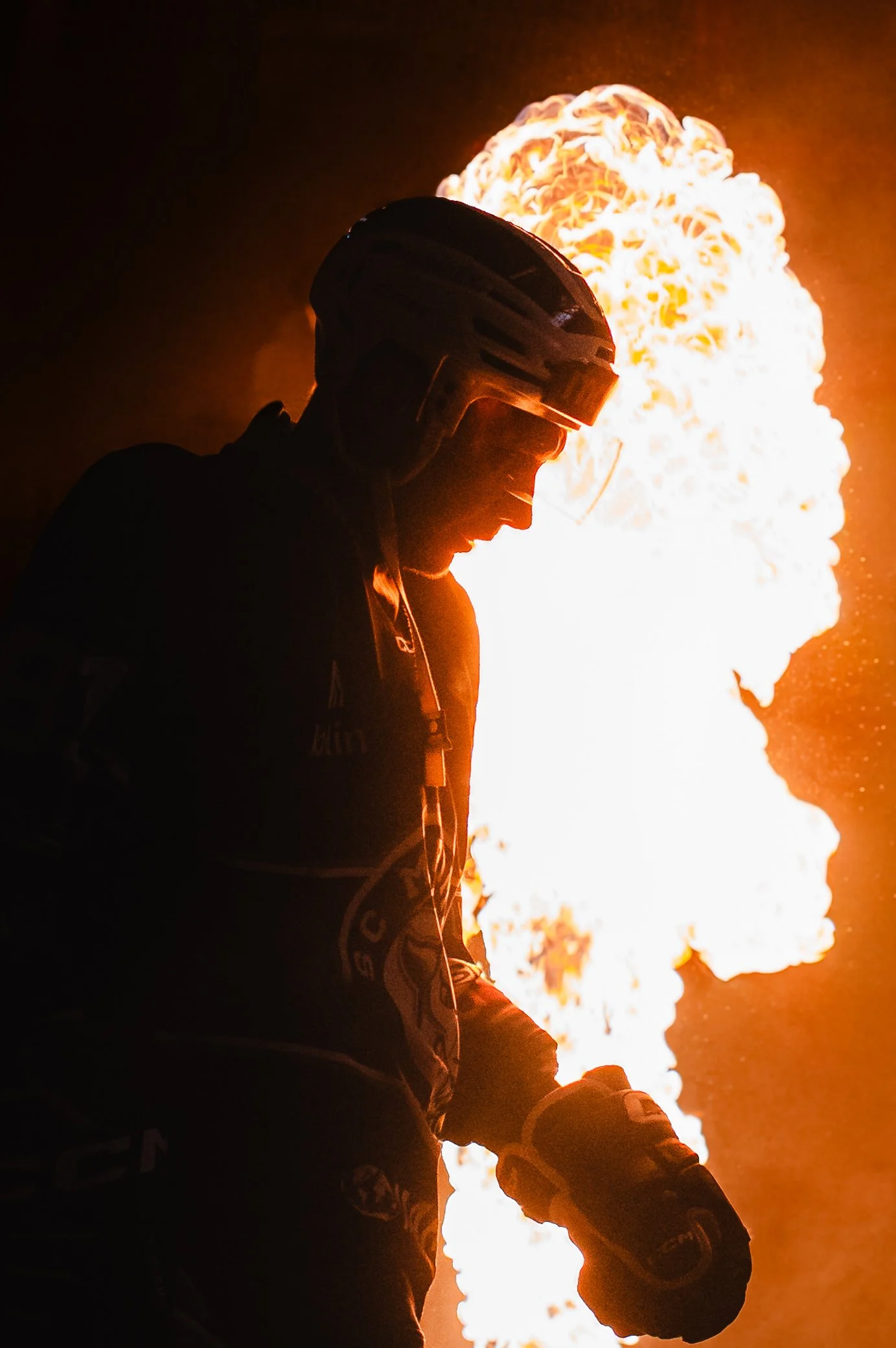 Silhouette of a person wearing a helmet and racing gear, standing in front of a large explosion or fire.