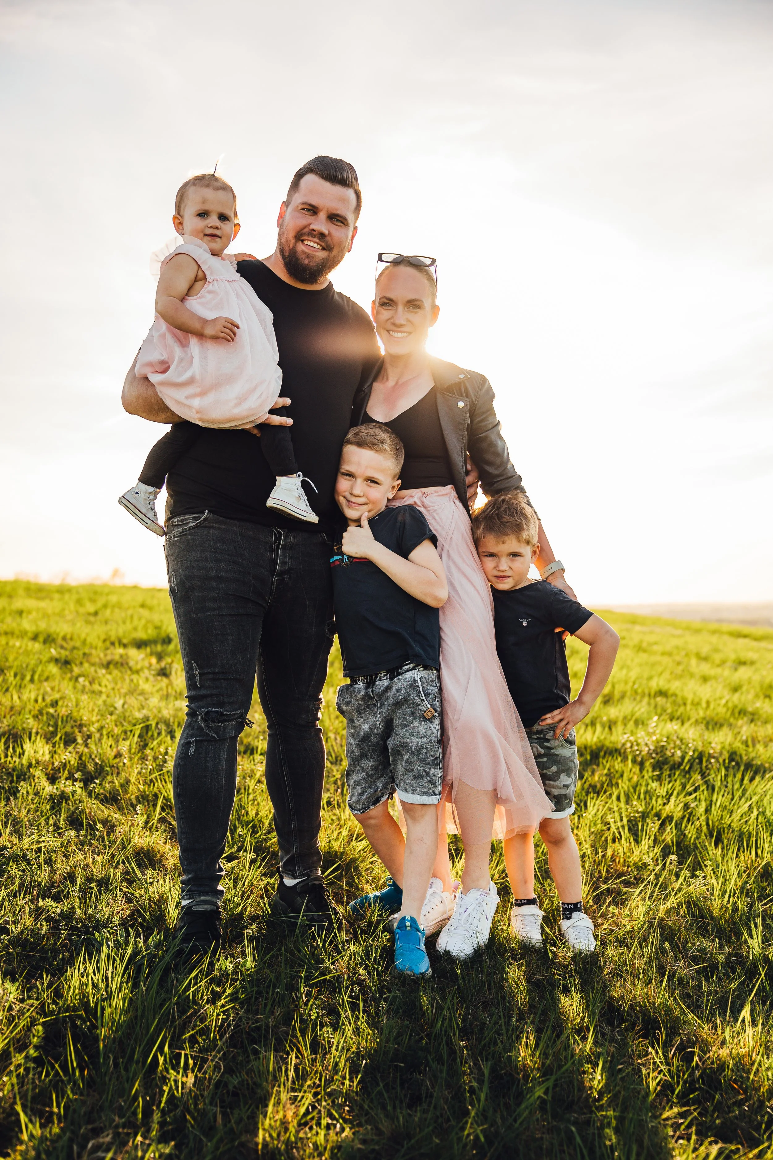 Family of five standing outdoors on grassy field at sunset, smiling at camera.