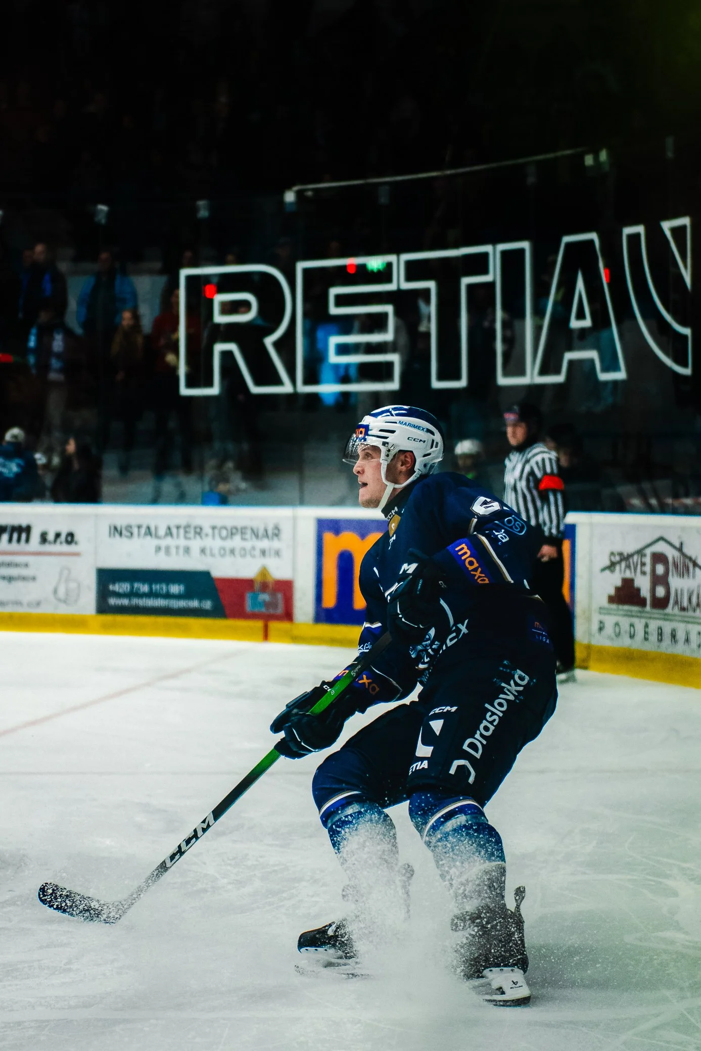 A hockey player in a blue uniform skating on ice with a hockey stick, wearing a helmet, in an indoor ice rink with a large illuminated 'RESTAURANT' sign in the background.
