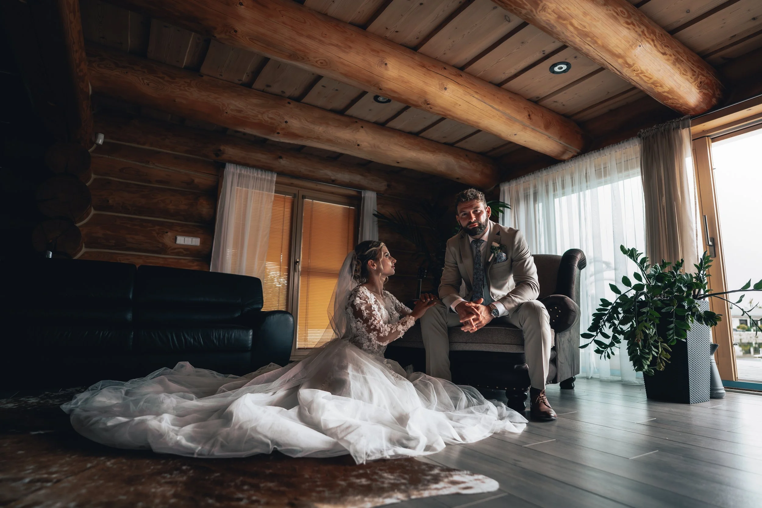 A bride in a wedding dress sitting on the floor and holding hands with a groom in a suit, sitting on a sofa in a cozy, wood-paneled room with large windows and curtains.