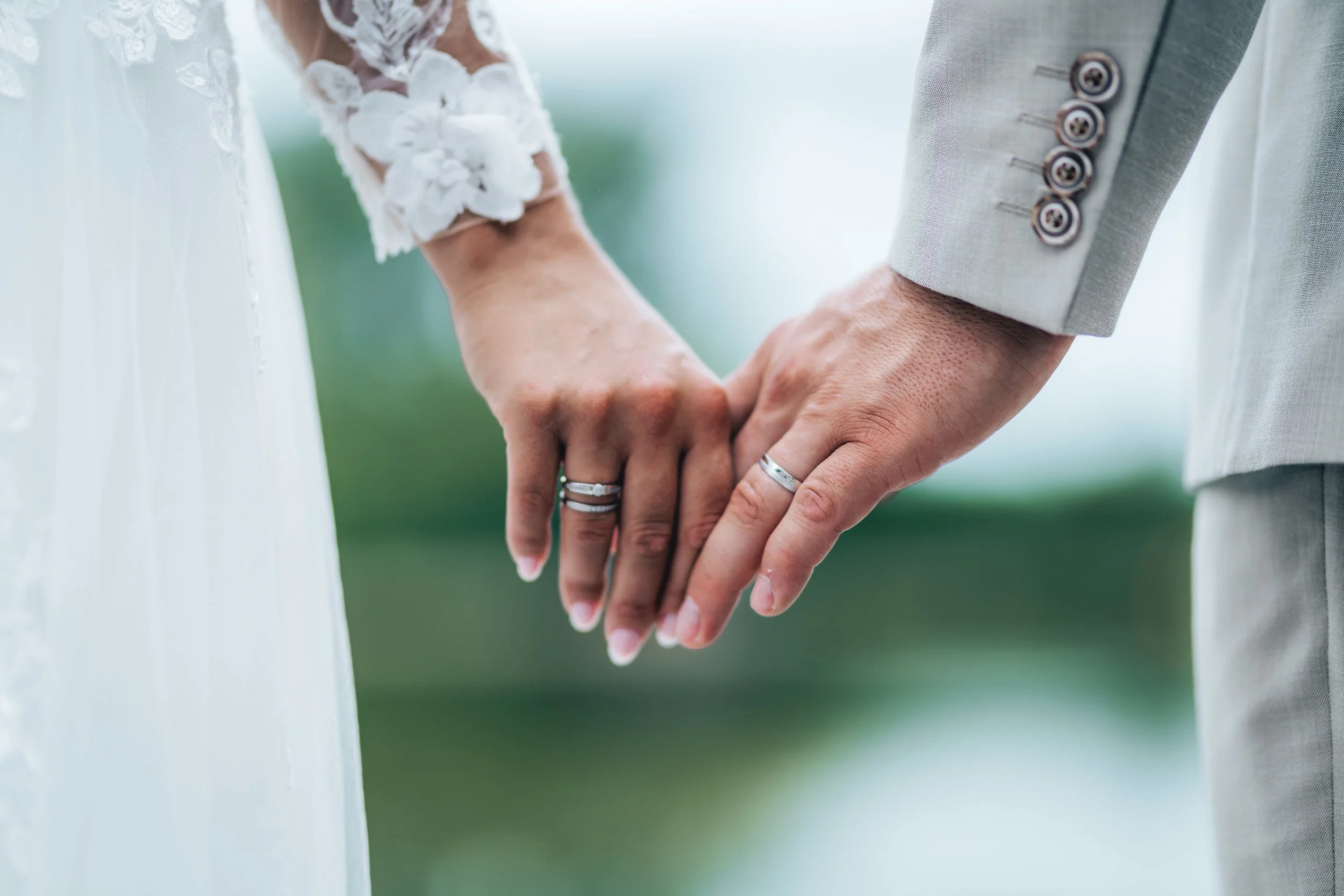 Close-up of a bride and groom holding hands, showing wedding rings, with the bride wearing a lace dress and the groom in a light-colored suit.