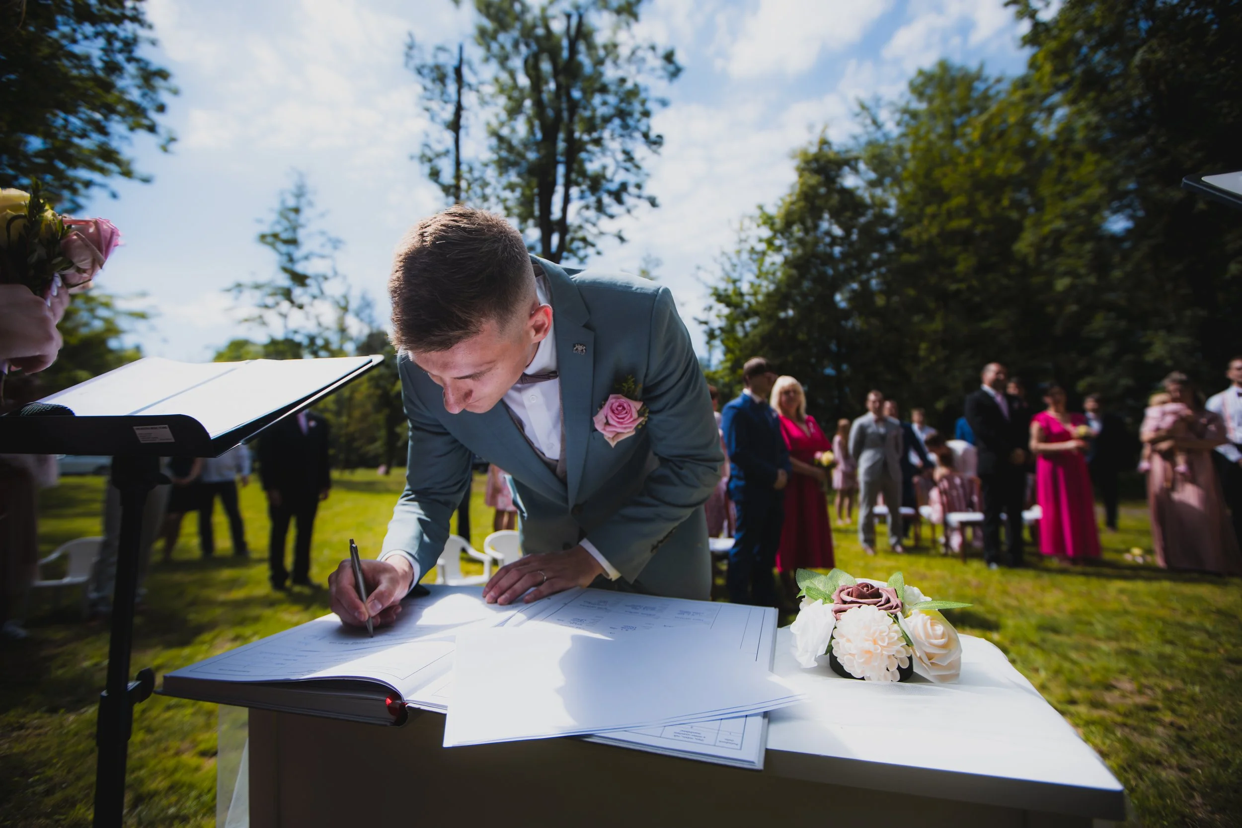 Groom signing a marriage register outdoors at a wedding ceremony with guests in the background.