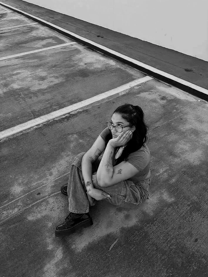 A young woman with glasses, tattoos, and dark hair tied in a high ponytail, sitting on the ground in an empty parking lot with her head resting on her hand, looking to her right.
