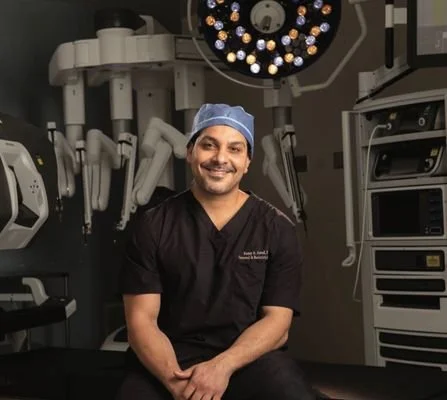 A male healthcare professional wearing scrubs and a surgical cap, sitting in an operating room with medical equipment and surgical lights overhead.