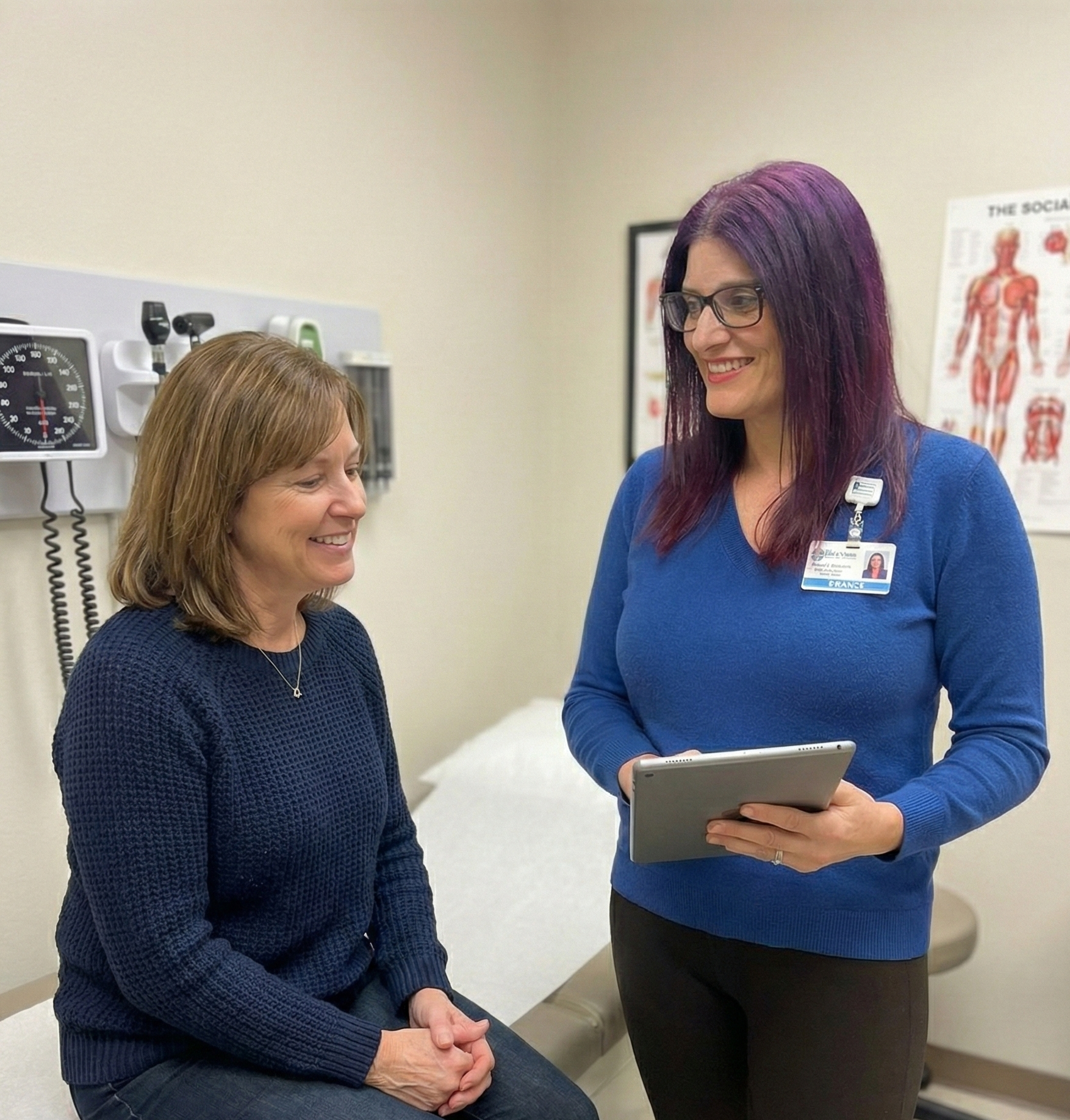 A healthcare professional with purple hair, glasses, and a blue uniform is talking to a seated woman with shoulder-length brown hair, a navy sweater, and jeans in a medical office. The professional is holding a tablet and has a name badge. A medical chart and a blood pressure monitor are visible in the background.