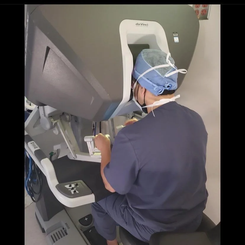A medical professional operating a da Vinci robotic surgical system, dressed in scrubs, a surgical cap, and mask, in a hospital or operating room environment.