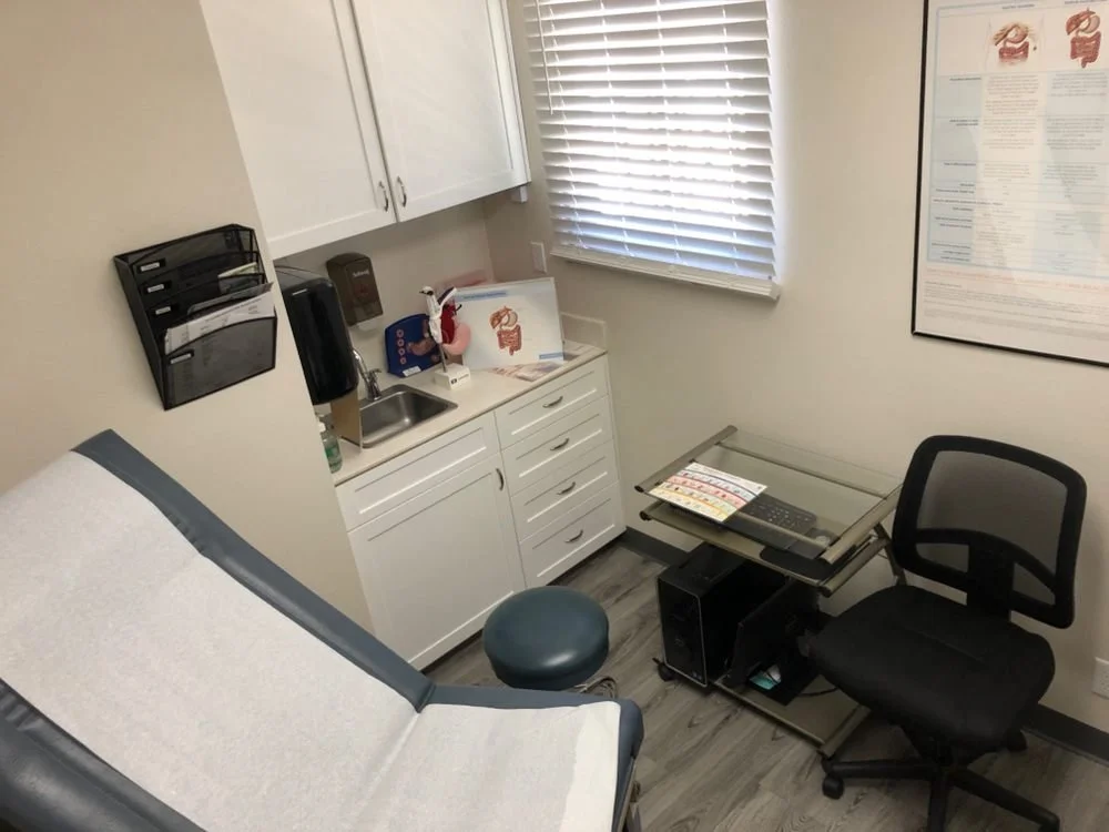 Medical examination room with a patient chair covered with white paper, a small sink area, and a desk with a computer, paperwork, and a chair. There is a window with blinds and a large chart on the wall.